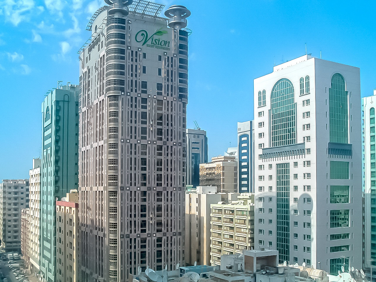 The image depicts a cityscape with several tall buildings. The tallest building in the center has the word 'Vision' on it, indicating it might be an office or commercial building. The architecture varies, with modern glass facades and more traditional designs. The sky is clear, suggesting a bright and sunny day. The buildings are closely packed, typical of an urban environment.