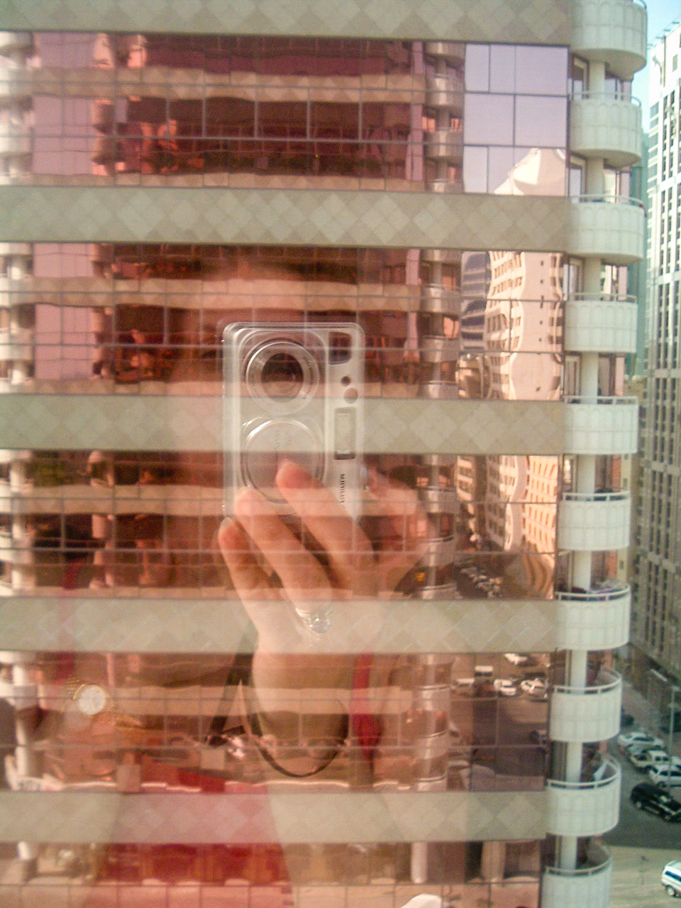 The image shows a reflection in a glass window. The reflection captures a person taking a photo with a digital camera. The background reveals a modern building with multiple floors and large windows. The photo is slightly blurred, making it difficult to discern finer details. The overall scene suggests an urban environment.