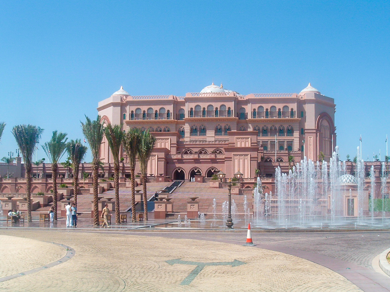 The image depicts a grand, ornate building with intricate architectural details, likely a palace or a luxurious hotel. The building features multiple levels, arched windows, and domed roofs. In front of the building, there is a large fountain with water jets, and the area is landscaped with palm trees and paved walkways. Several people are seen walking and sitting around the fountain, enjoying the sunny weather.