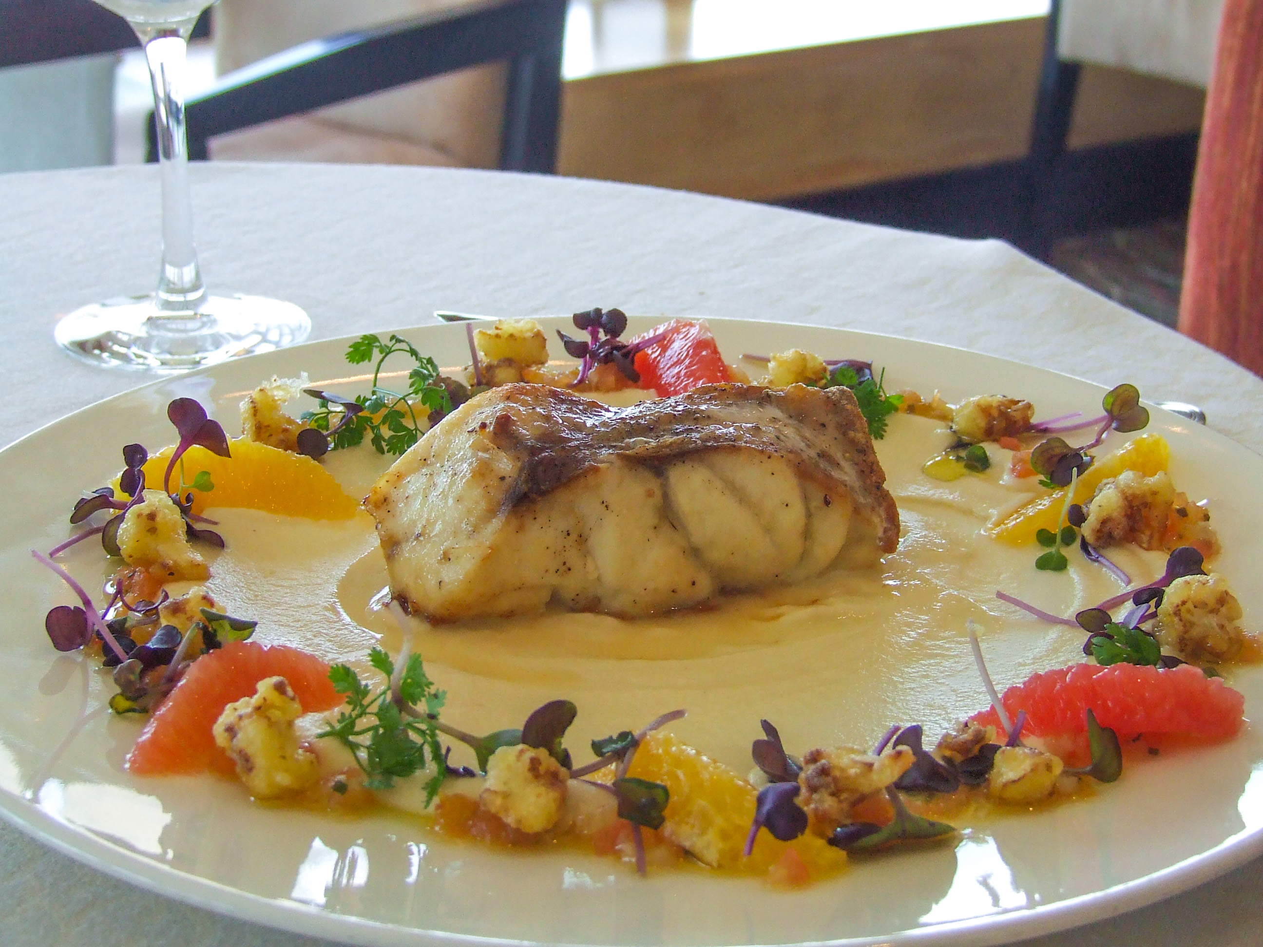 The image depicts a beautifully plated dish featuring a piece of fish as the main component, surrounded by a creamy sauce and an assortment of garnishes including microgreens, roasted vegetables, and herbs. The presentation is elegant, suggesting it is from a high-end restaurant.