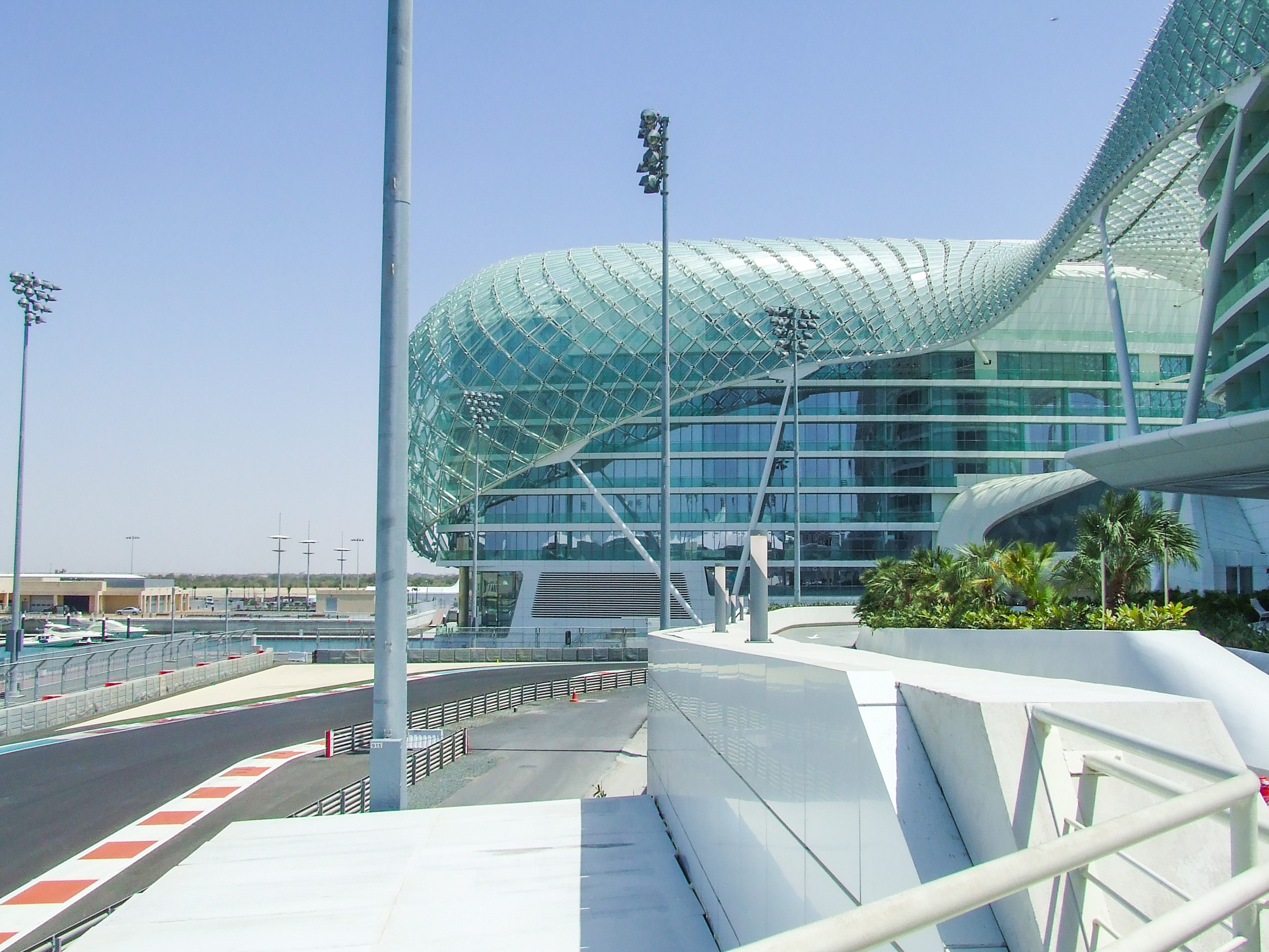The image depicts a modern architectural structure with a distinctive, curved glass facade, likely part of a large, contemporary building complex. The scene includes a racing track with red and white curbs, suggesting it might be a circuit or a track within the complex. The area is well-maintained with some greenery and palm trees, and the sky is clear, indicating good weather. The overall setting appears to be a blend of a high-tech facility and a racing venue, possibly an airport or a similar large-scale infrastructure.