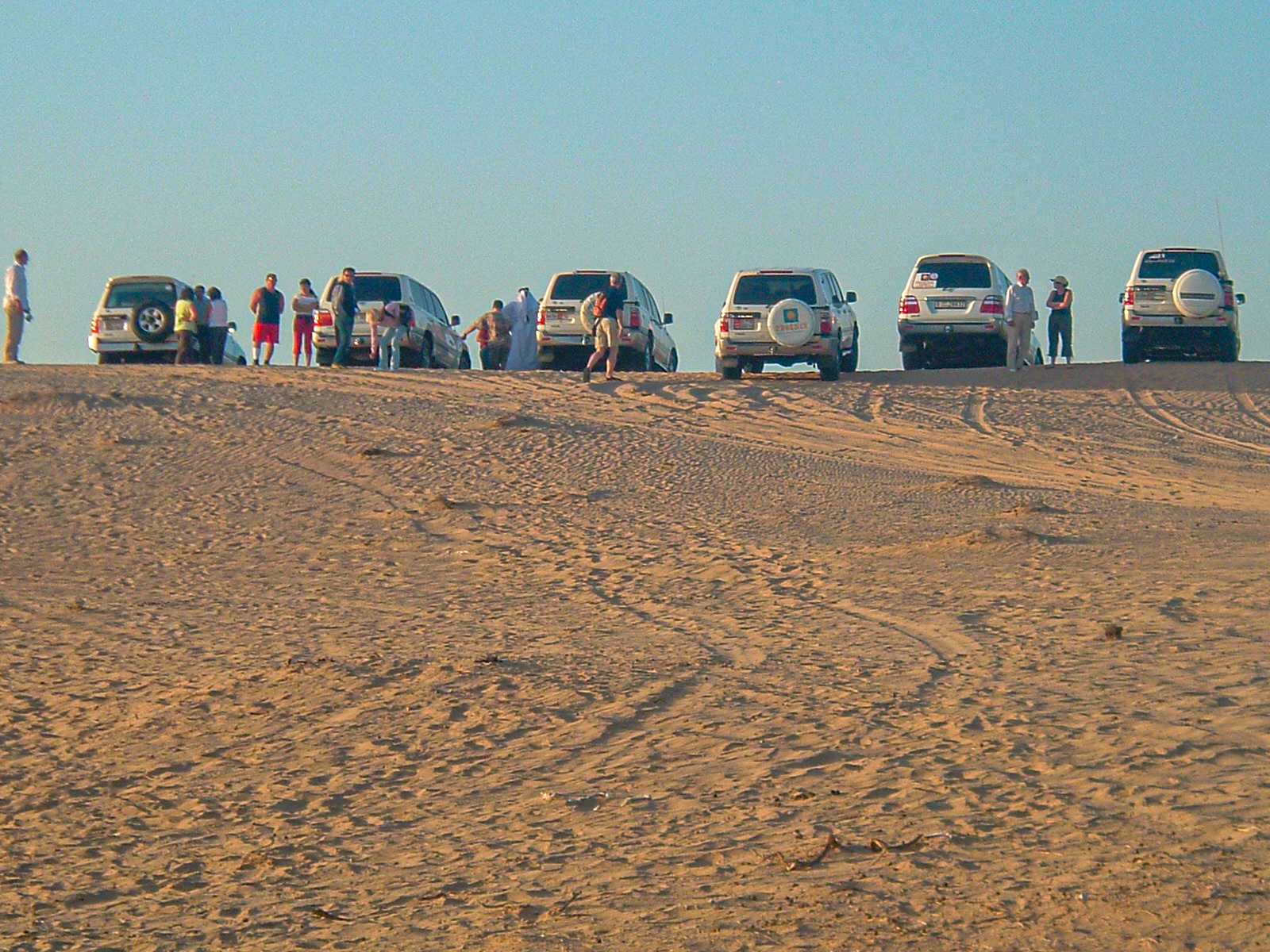 The image shows a group of people gathered around several off-road vehicles parked on a sandy terrain, likely participating in a desert excursion or adventure activity.
