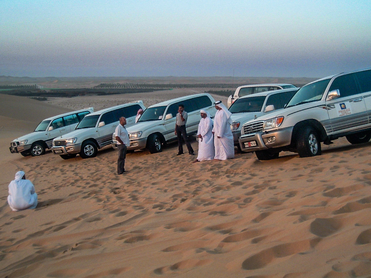 The image shows a group of people standing in a desert landscape with several vehicles parked on the sandy terrain. The individuals are dressed in traditional and Western attire, suggesting a mix of cultural backgrounds. The scene appears to be set in a desert environment, possibly during a desert safari or a similar outdoor activity.