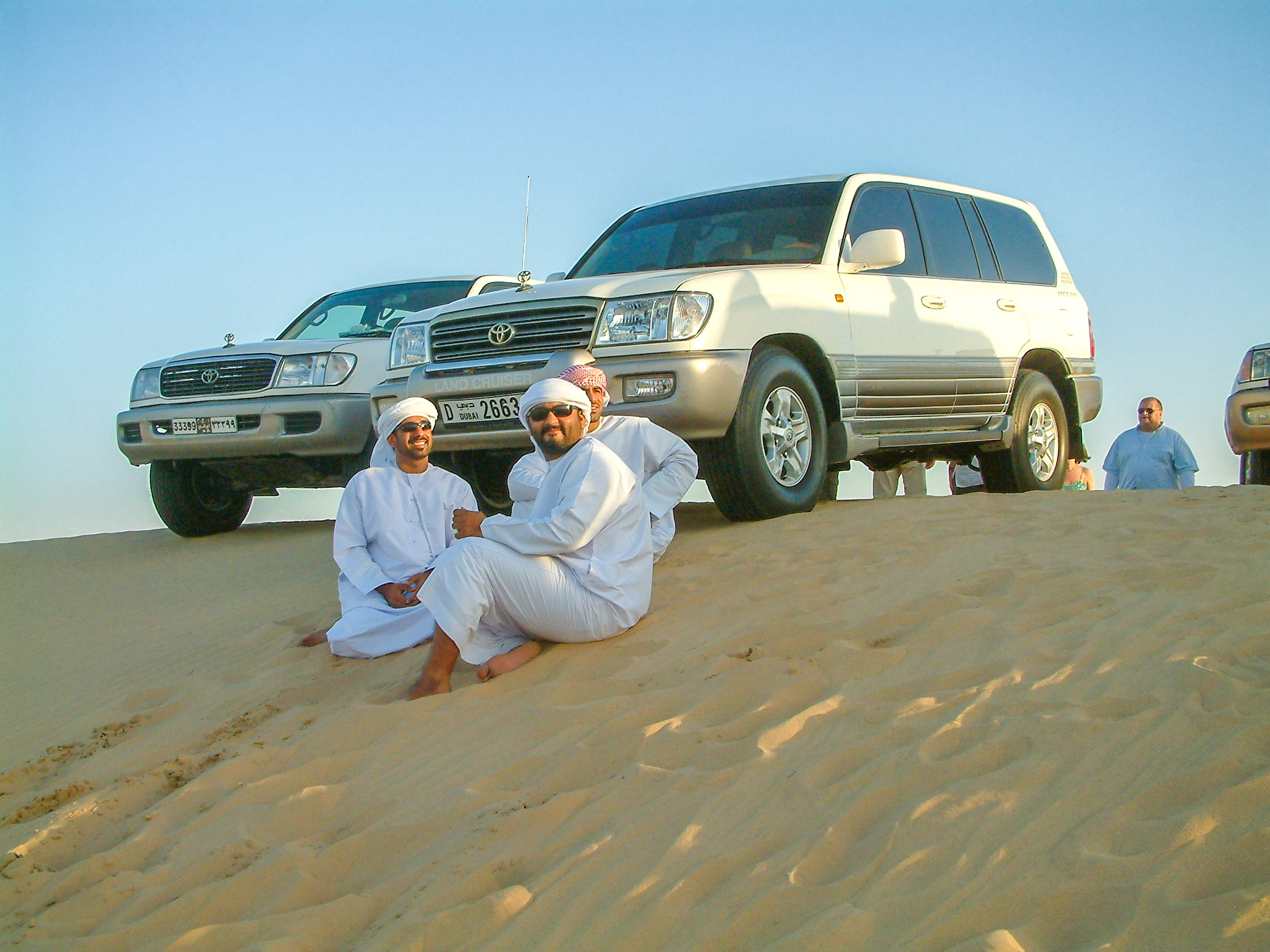 The image shows two men dressed in traditional white attire sitting on a sand dune with two Toyota SUVs parked behind them. The men appear to be enjoying their time in a desert setting, with clear skies and bright sunlight. Other individuals are visible in the background, suggesting a group outing or adventure.
