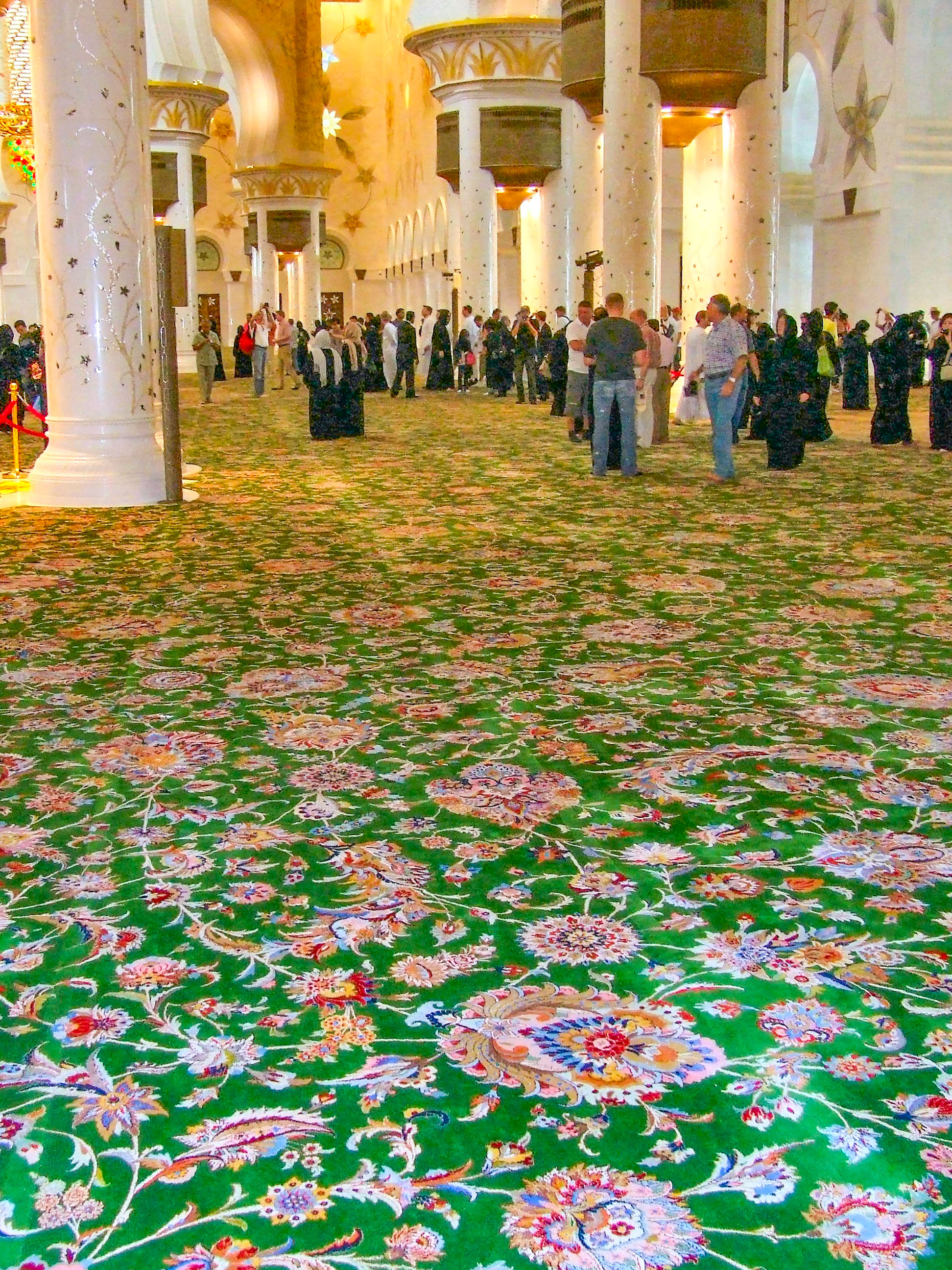 The image depicts a large, ornate interior space with high ceilings, grand columns, and intricate floral carpeting. The area is filled with people, some of whom appear to be tourists taking photos. The architecture features white columns and arches, with decorative elements and lighting that highlight the grandeur of the space. The overall atmosphere is one of awe and admiration for the detailed and luxurious design.
