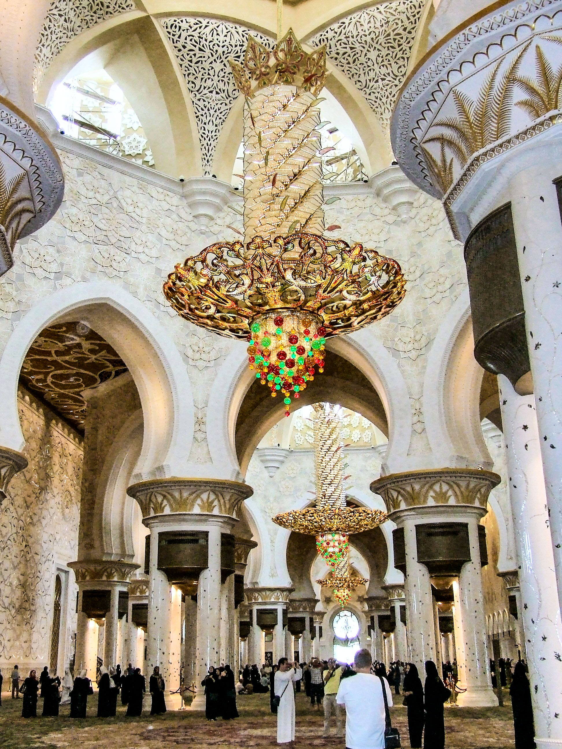 The image depicts the interior of the Sheikh Zayed Mosque in Abu Dhabi, showcasing its grand architectural features such as large white marble columns, intricate arches, and ornate chandeliers. The mosque's design includes detailed Islamic patterns and calligraphy on the walls and ceilings. The space is filled with visitors, some of whom are taking photos and observing the surroundings. The overall atmosphere is one of awe and admiration for the mosque's beauty and scale.