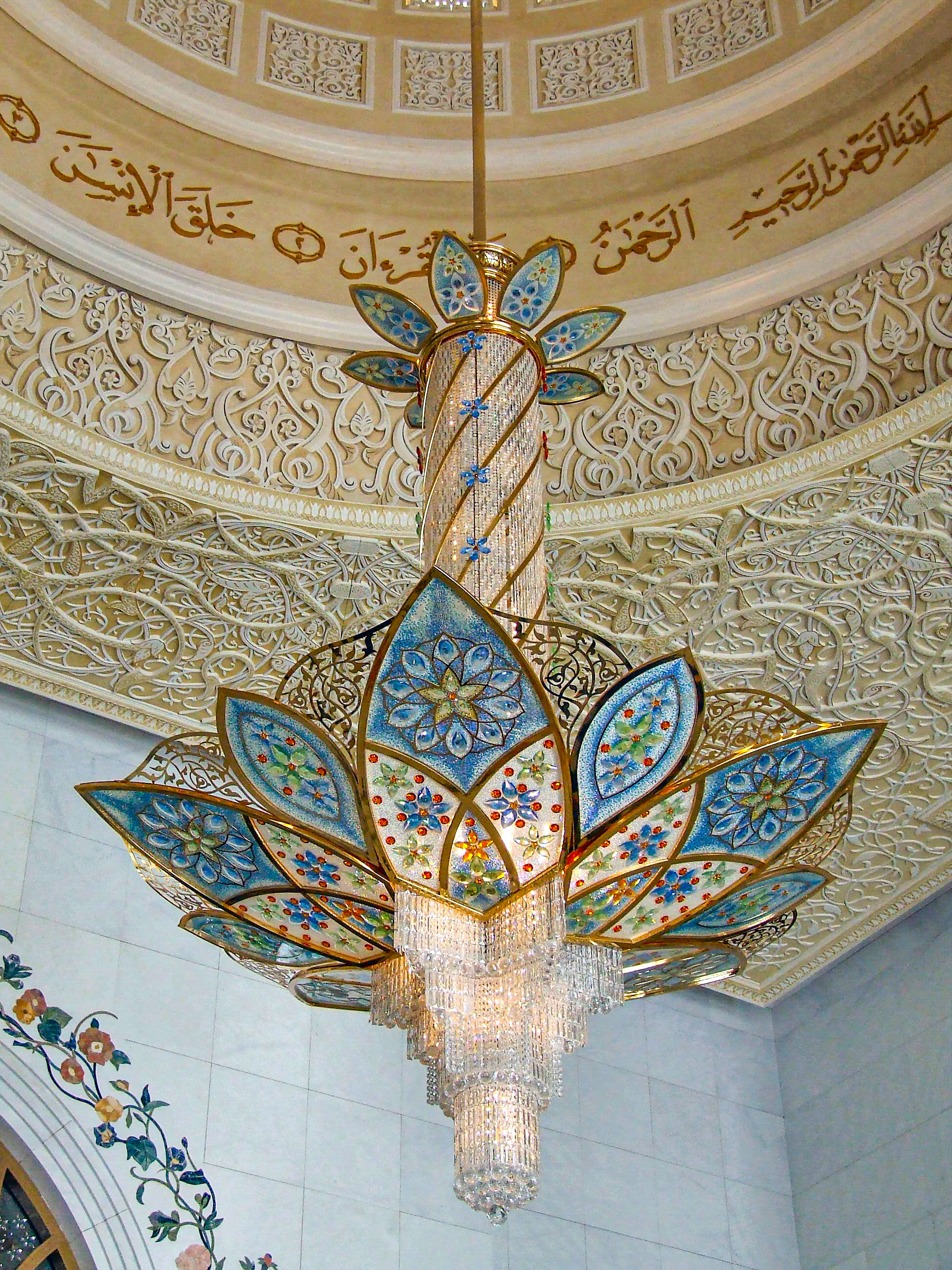 The image depicts an intricately designed chandelier hanging from a ceiling adorned with detailed Islamic art and calligraphy. The chandelier features a floral design with colorful, mosaic-like patterns and crystal embellishments. The ceiling showcases elaborate carvings and Arabic script, creating a rich and ornate visual effect.
