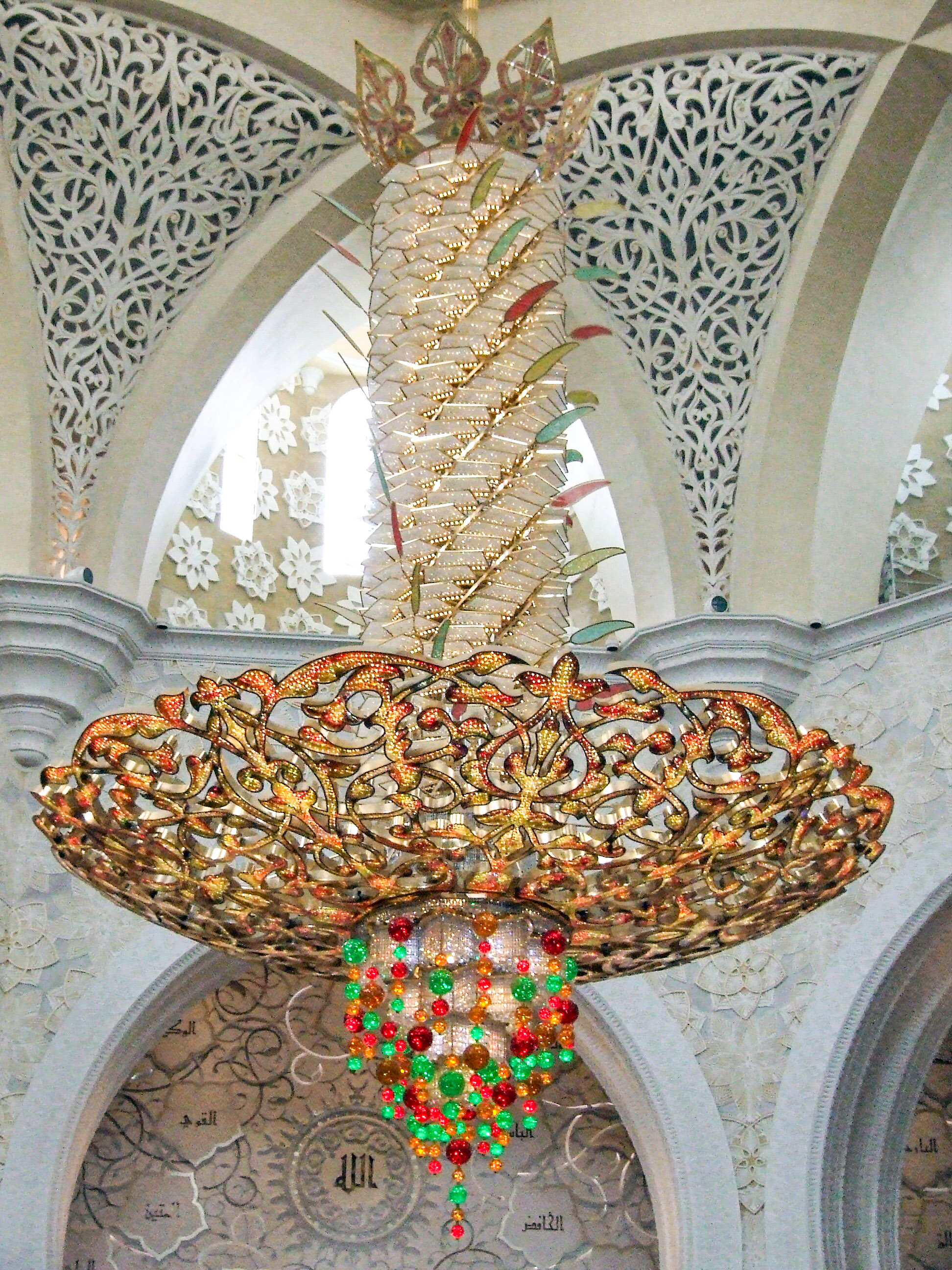 The image showcases an ornate chandelier hanging from an intricately designed ceiling. The chandelier features elaborate patterns and is adorned with colorful beads and embellishments. The ceiling above has detailed carvings and floral motifs, adding to the overall grandeur and elegance of the setting.