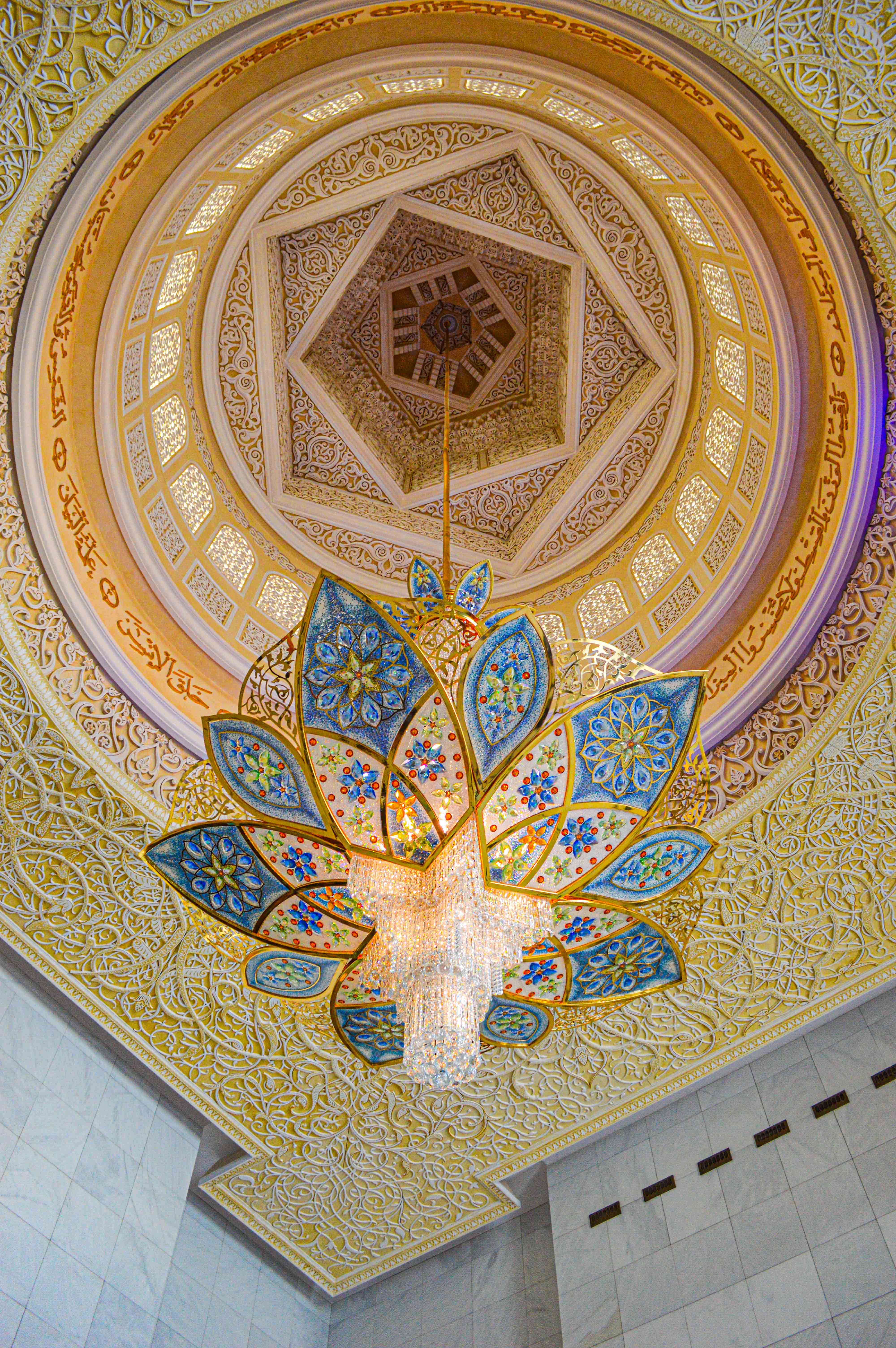 The image showcases an intricately designed ceiling featuring a large, ornate chandelier. The ceiling is adorned with detailed patterns and floral motifs, predominantly in gold and blue hues. The design includes geometric shapes and Arabic calligraphy, creating a visually stunning and symmetrical composition.