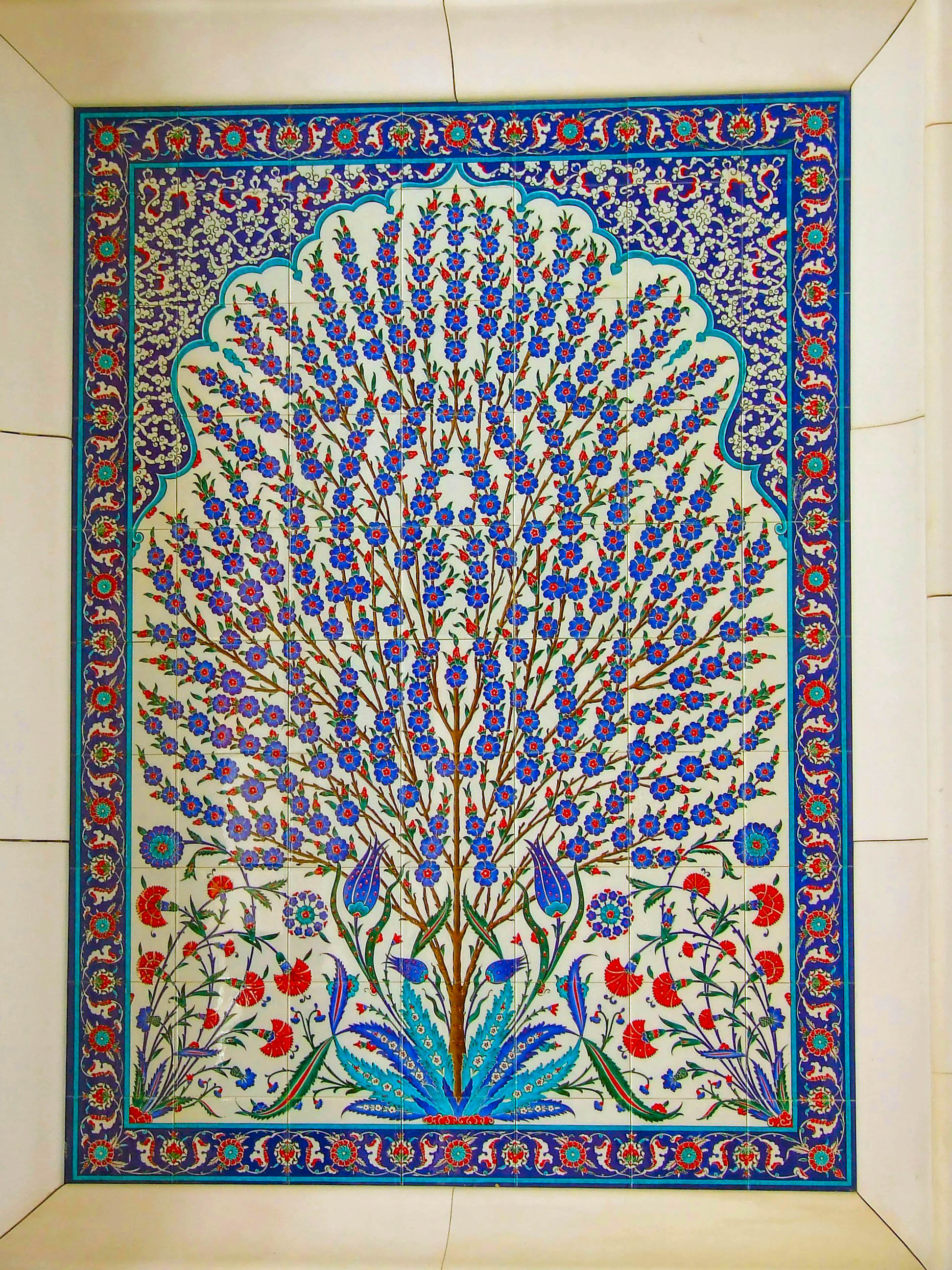 The image depicts a beautifully intricate tile artwork featuring a central tree with blue flowers and red accents, surrounded by detailed floral patterns and ornate borders. The design is framed with a blue and white border adorned with red and green floral motifs. The artwork showcases a harmonious blend of colors and patterns, typical of traditional Islamic art.