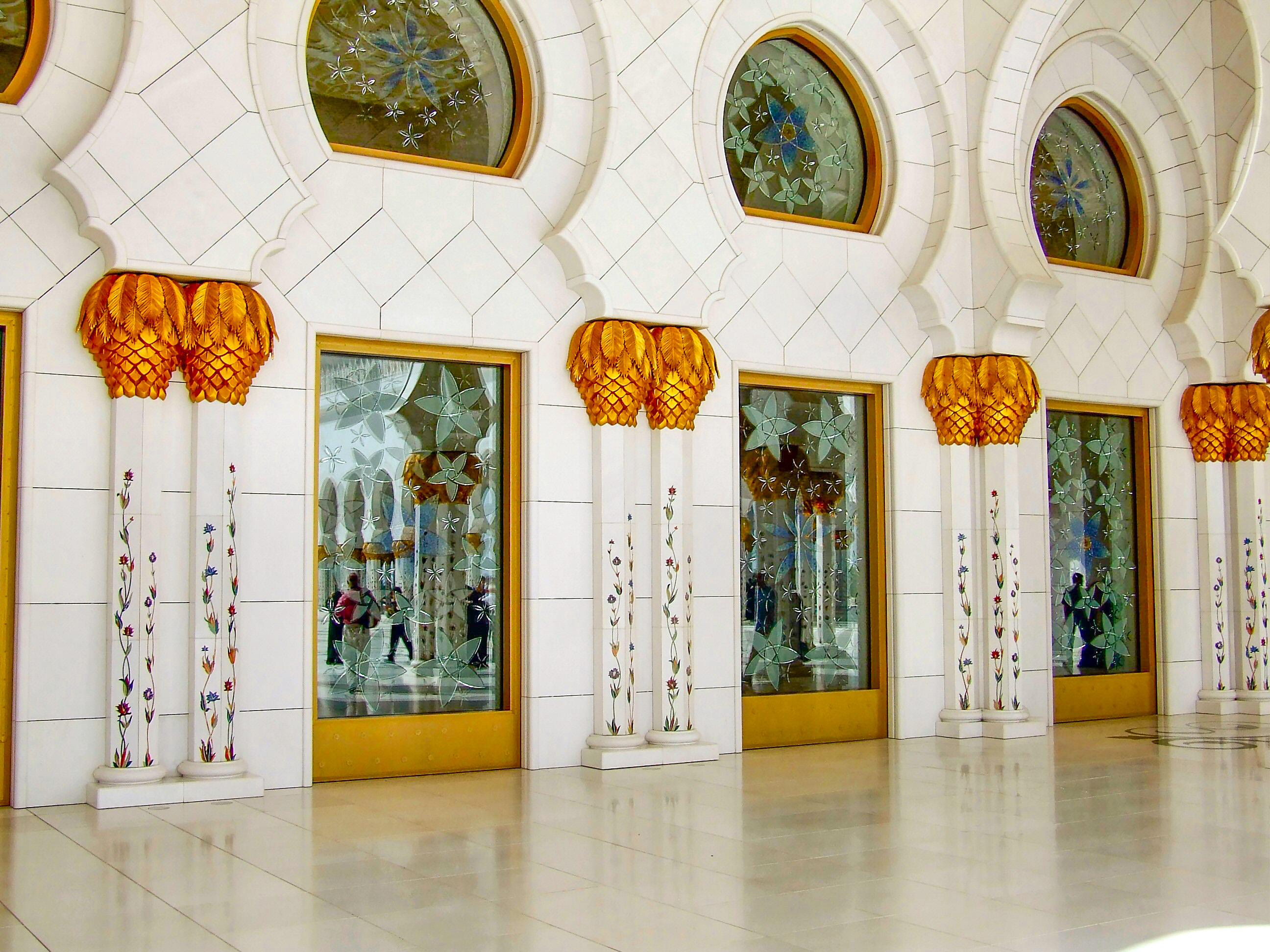 The image depicts an interior corridor with ornate architectural details. The walls are adorned with white tiles and feature large, intricately designed windows with stained glass. Each window is framed by gold-colored wood. Between the windows, there are tall, slender columns with colorful floral designs. Above each column, there are decorative light fixtures shaped like flowers with gold and orange hues.