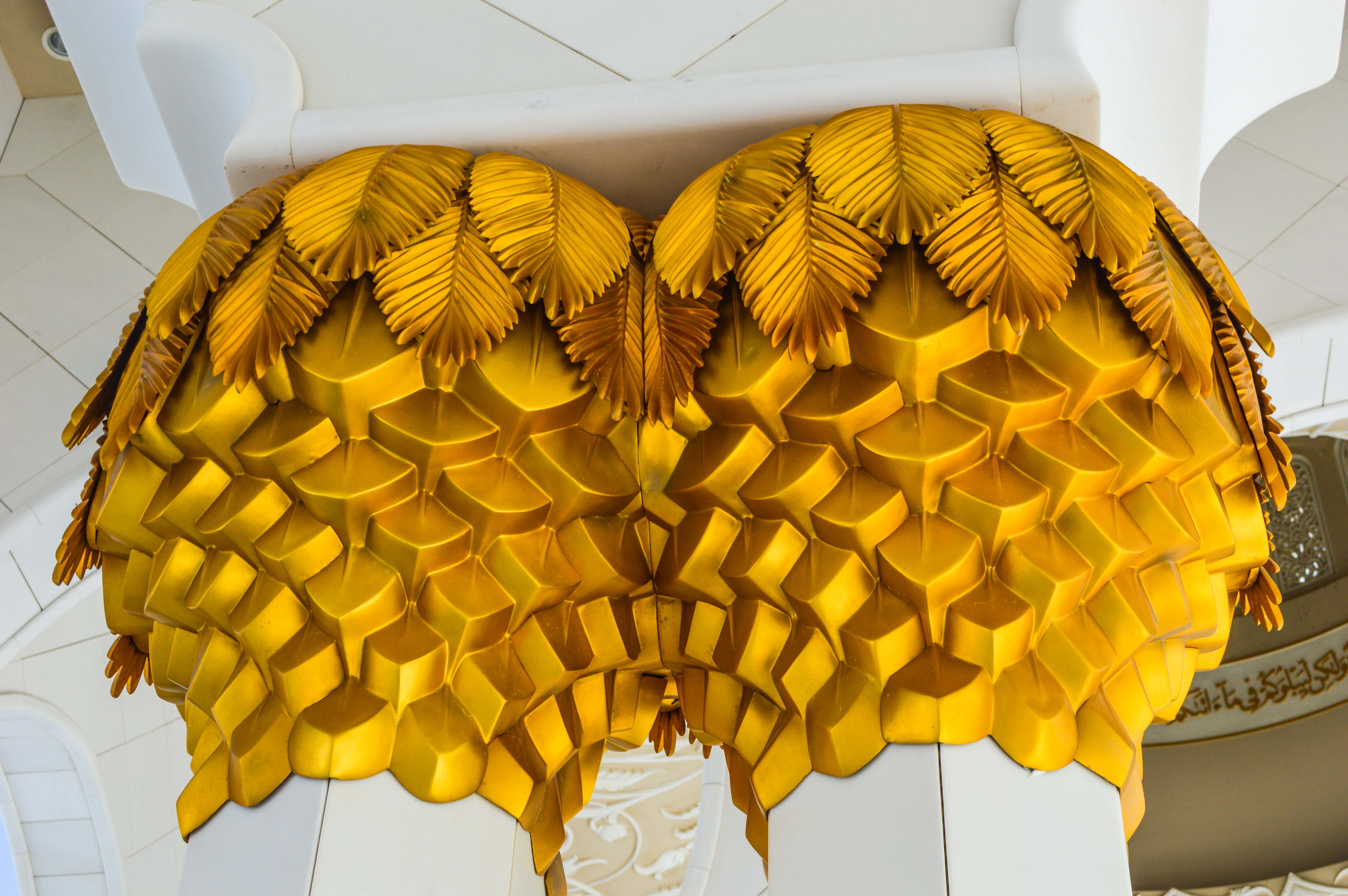The image showcases an intricately designed, large, yellow decorative piece hanging from a white ceiling. The piece features a combination of geometric and organic shapes, resembling a stylized, abstract representation of a bird or a similar creature. The design incorporates a series of interconnected, overlapping elements that create a three-dimensional effect. The background includes white, ornately decorated walls with Arabic calligraphy, suggesting that the location might be a mosque or an Islamic cultural site.
