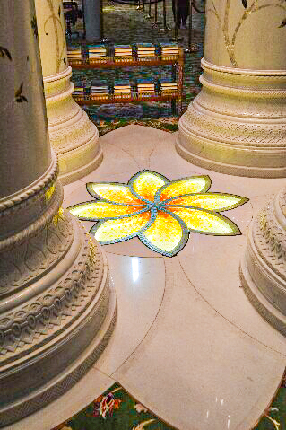 The image depicts an interior scene with ornate columns and a stained glass flower design on the floor. The columns are intricately decorated with patterns and the stained glass flower, featuring vibrant yellow and green colors, is positioned centrally between the columns. The floor is marble, adding to the elegance of the setting. In the background, there are rows of seats, suggesting that this could be a place of worship or a similar public building.