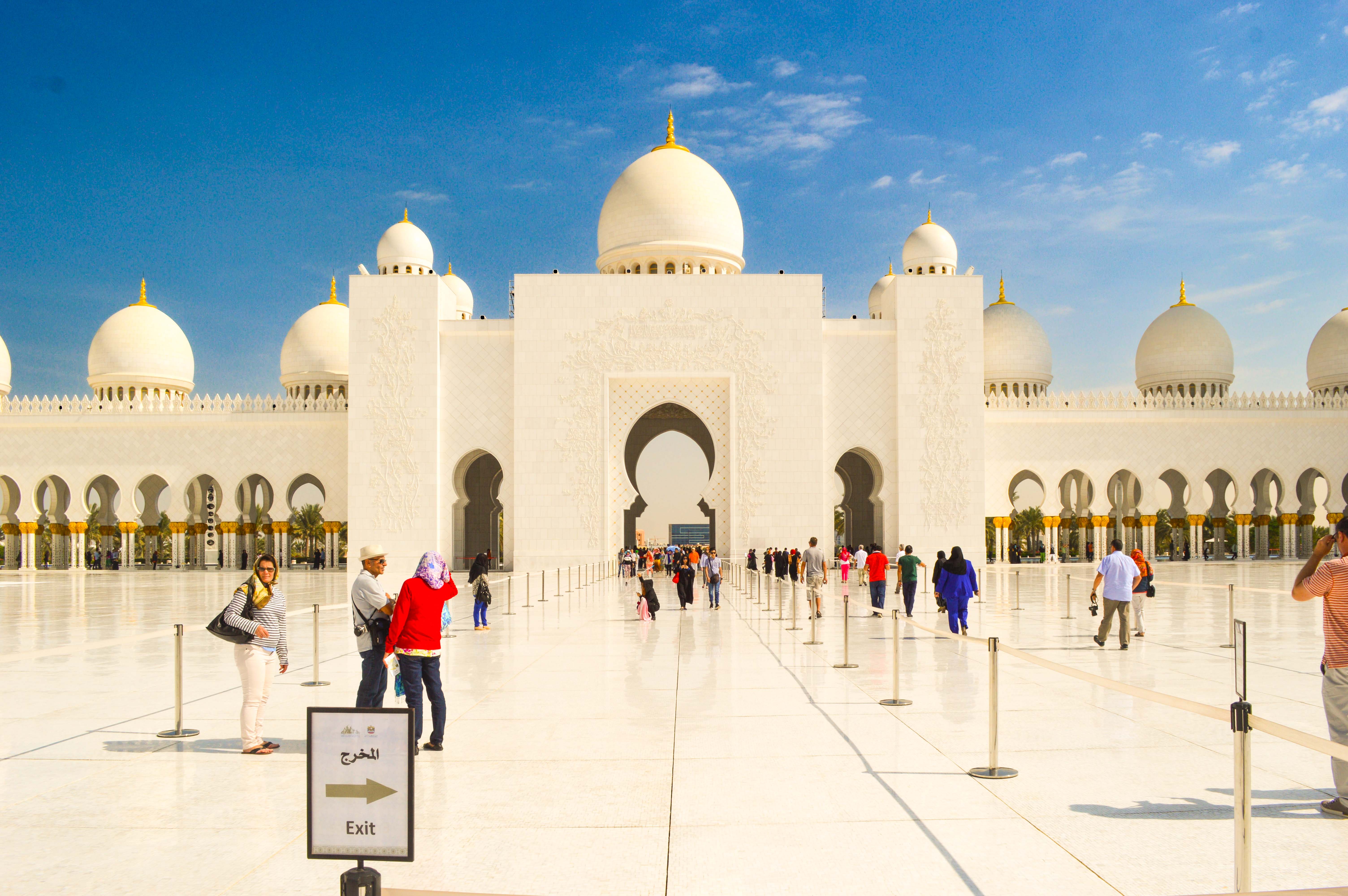 The image depicts the Sheikh Zayed Mosque in Abu Dhabi, UAE. It showcases the mosque's grand architectural features, including its large white domes, towering minarets, and intricate designs. The courtyard is spacious with marble flooring and is lined with arches. Visitors can be seen walking around and taking photographs. The sky is clear with a few scattered clouds, indicating a bright and sunny day.