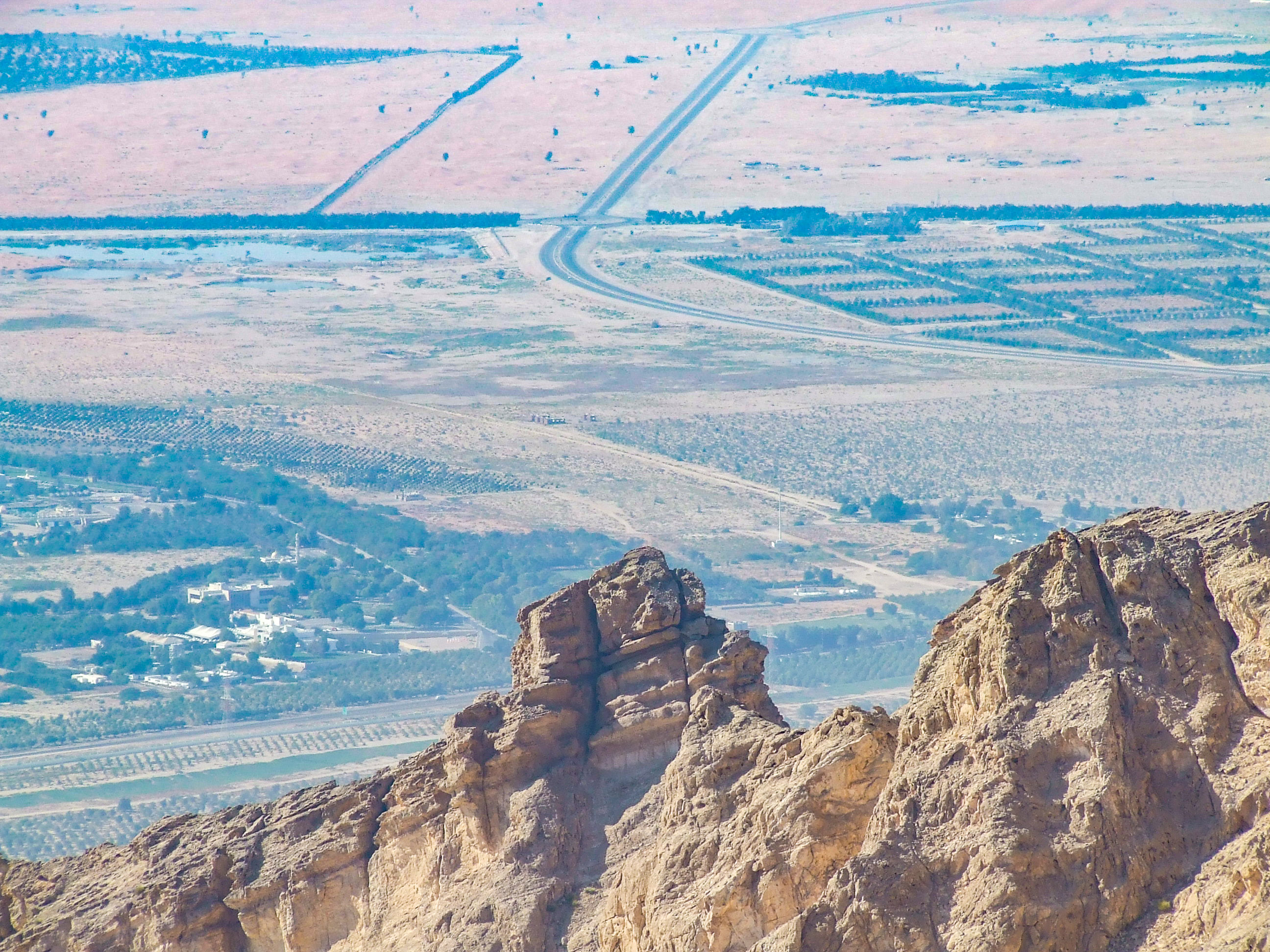 The image depicts a vast, arid landscape with a winding road cutting through the center. The foreground features rugged, rocky cliffs, while the background shows a patchwork of fields and sparse vegetation. The scene is characterized by its expansive, open terrain and the clear, winding road that stretches into the distance.