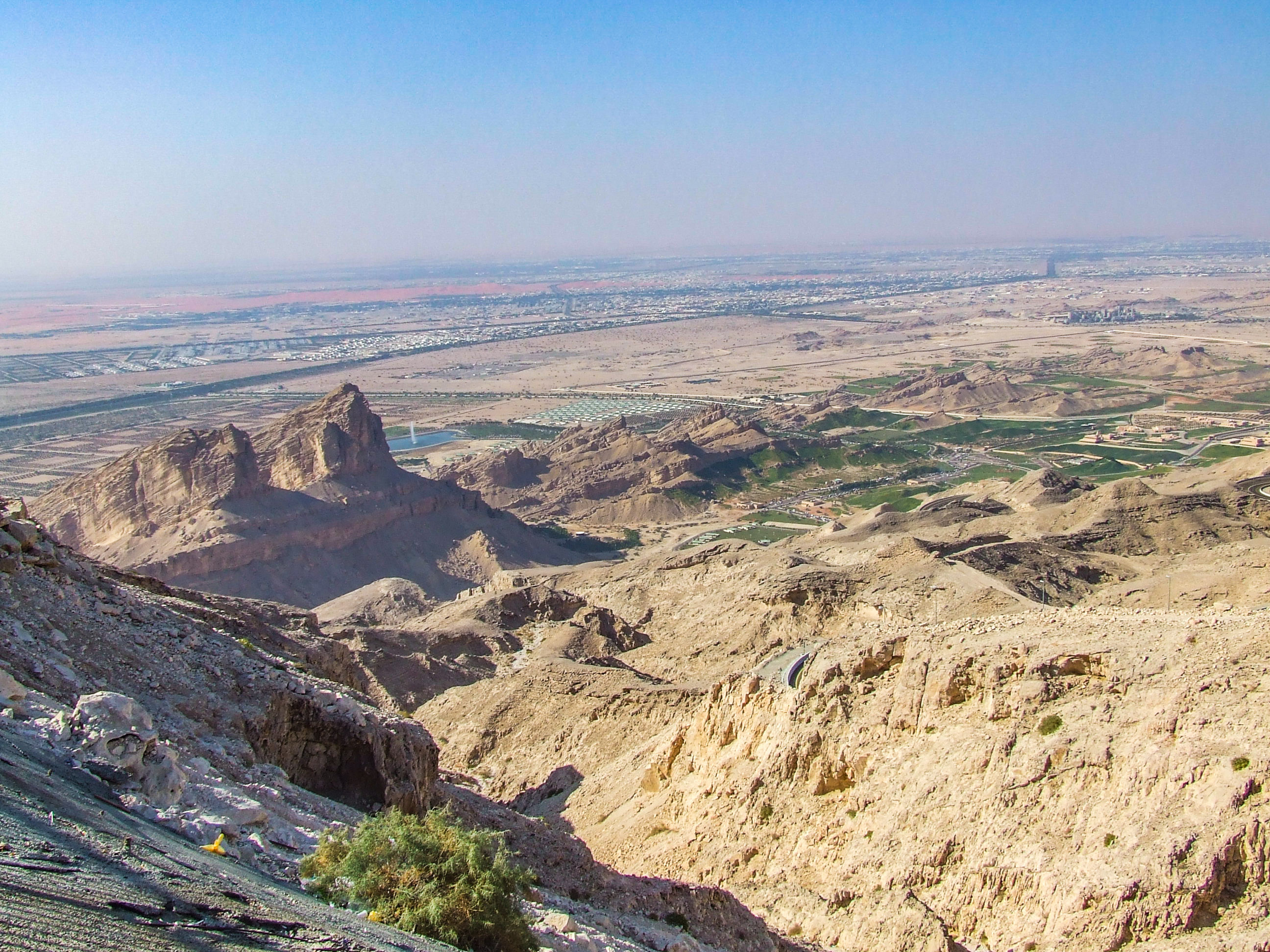 The image depicts a vast, arid landscape with rugged terrain and rocky formations. In the foreground, there are steep cliffs and jagged peaks. The midground features a series of hills and valleys with sparse vegetation. In the distance, a large urban area stretches across the horizon under a clear sky.
