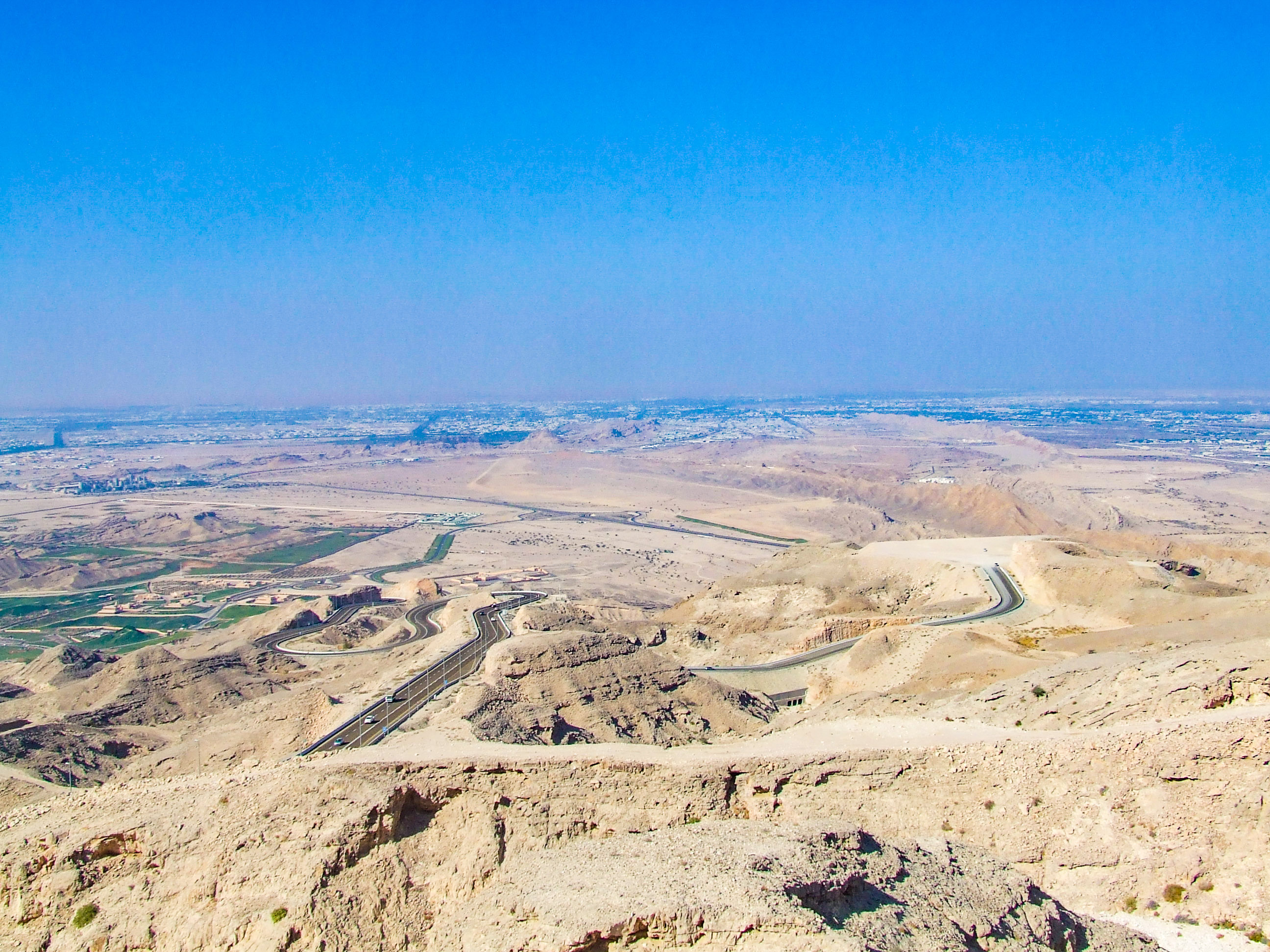 The image depicts a vast desert landscape with winding roads and sparse vegetation. The terrain is mostly barren with some green patches, likely irrigated areas. The sky is clear, and the overall scene suggests a remote, arid region with minimal human habitation.