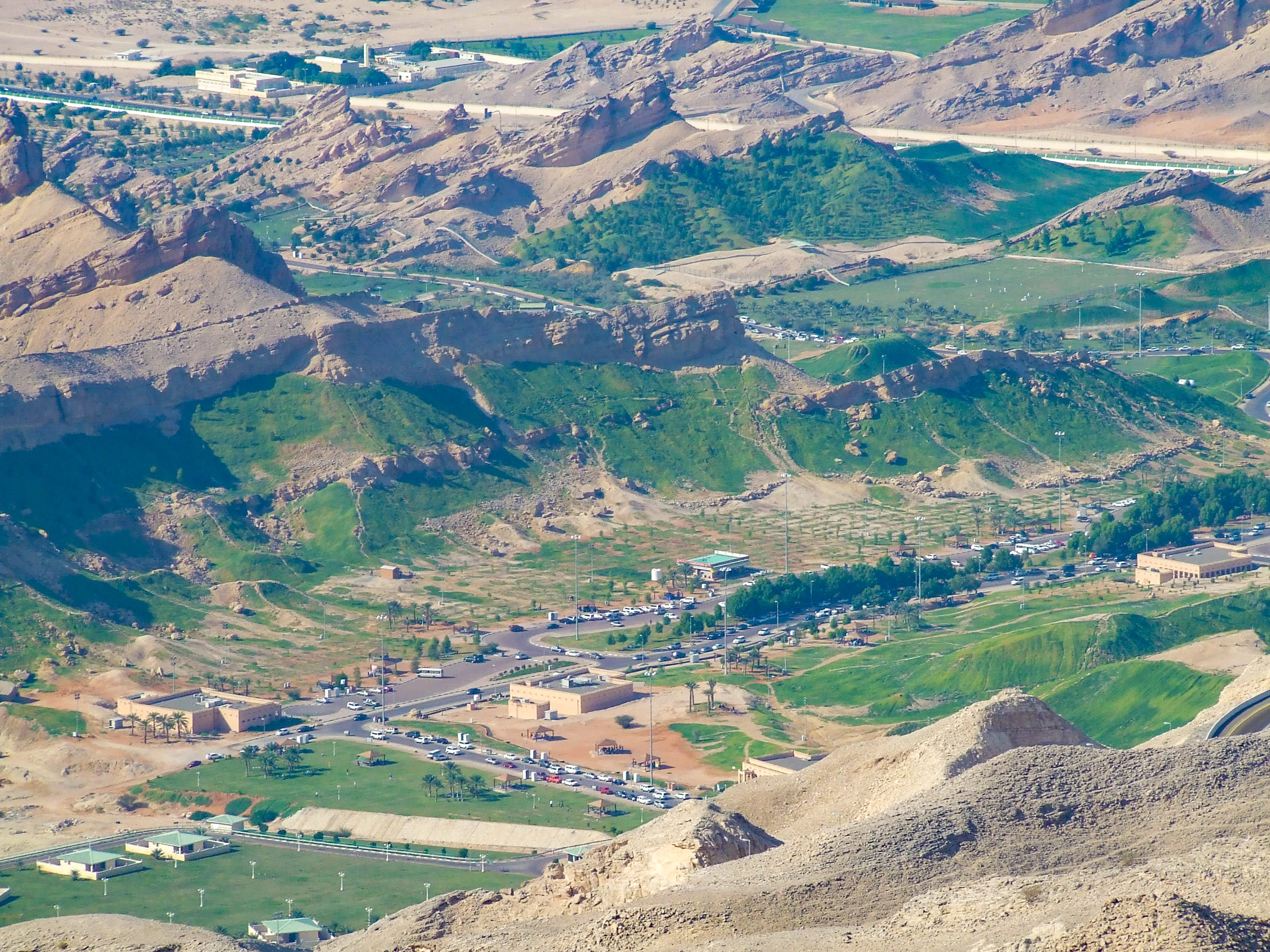 The image depicts a hilly and arid landscape with a small settlement nestled within it. The area features a mix of green patches and rocky terrain, with buildings, roads, and vehicles visible. The settlement appears to be surrounded by rugged hills and mountains, creating a scenic and isolated environment.