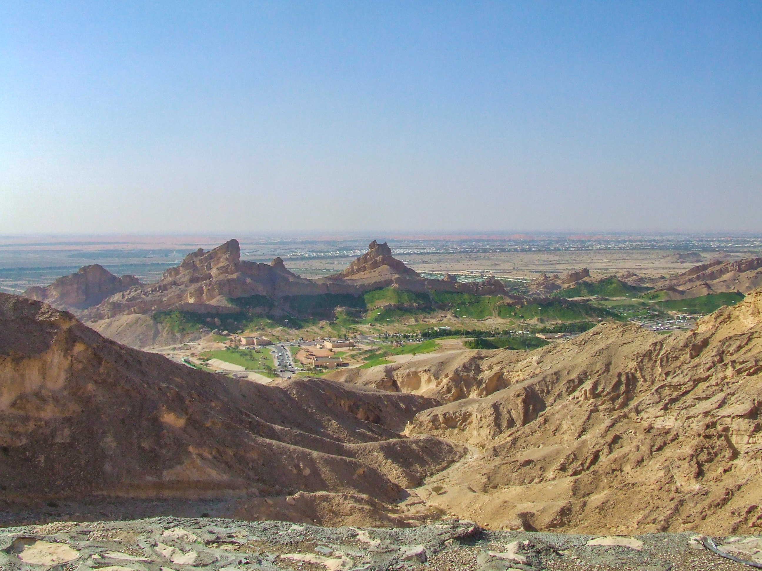 The image depicts a rugged, arid landscape with rocky formations and sparse vegetation. In the foreground, there are steep, barren hills with a small settlement or community visible in the mid-ground. The background features more rocky outcrops and a distant, flat expanse that stretches to the horizon under a clear sky.