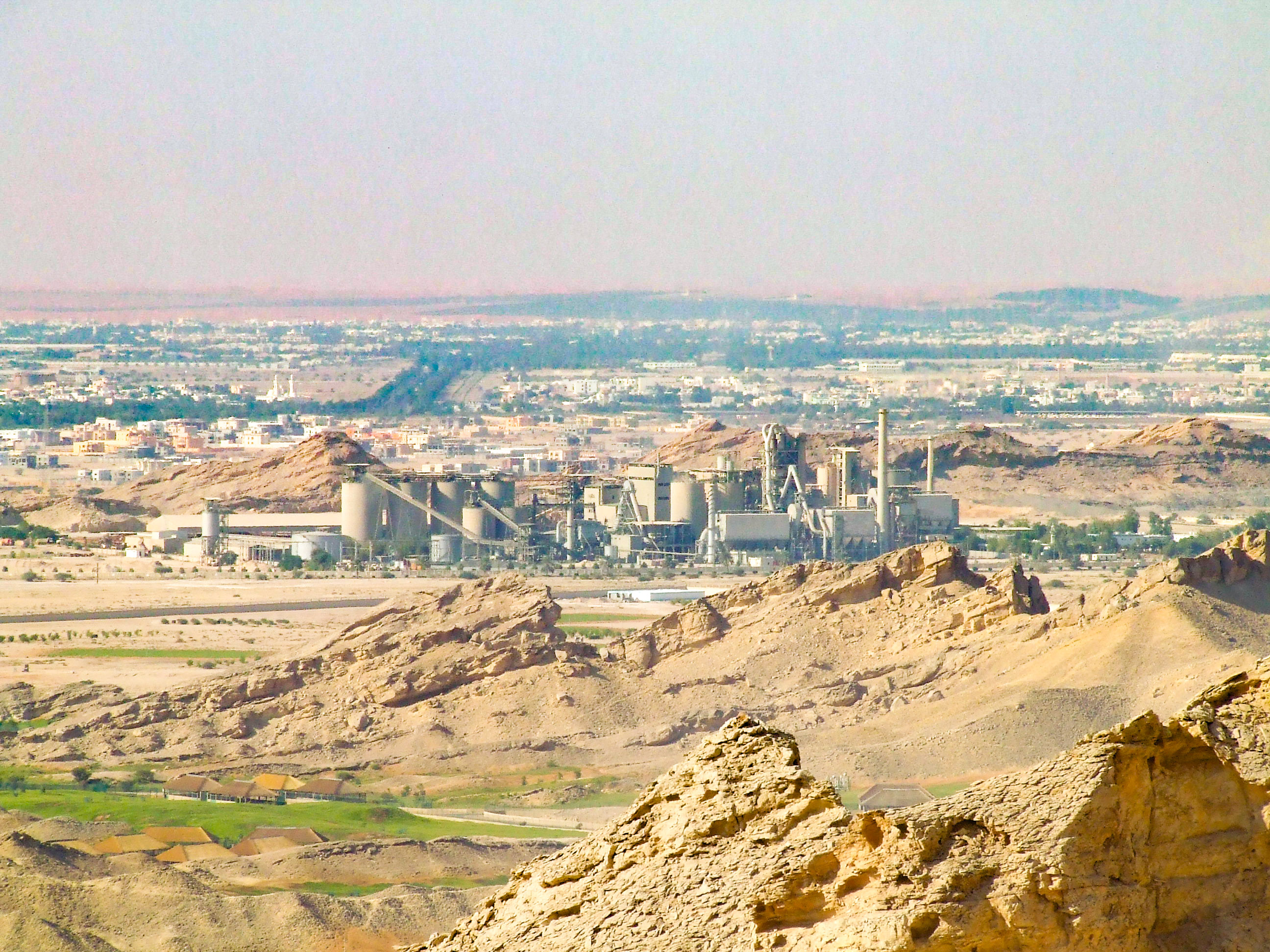 The image depicts an industrial area with a large factory or plant situated in a desert-like environment. The factory is surrounded by barren, rocky terrain with sparse vegetation. In the background, there is an expansive urban area with numerous buildings and infrastructure stretching towards the horizon. The sky is hazy, suggesting a possibly arid or polluted atmosphere. The overall scene suggests a juxtaposition of industrial development against a natural, rugged landscape.