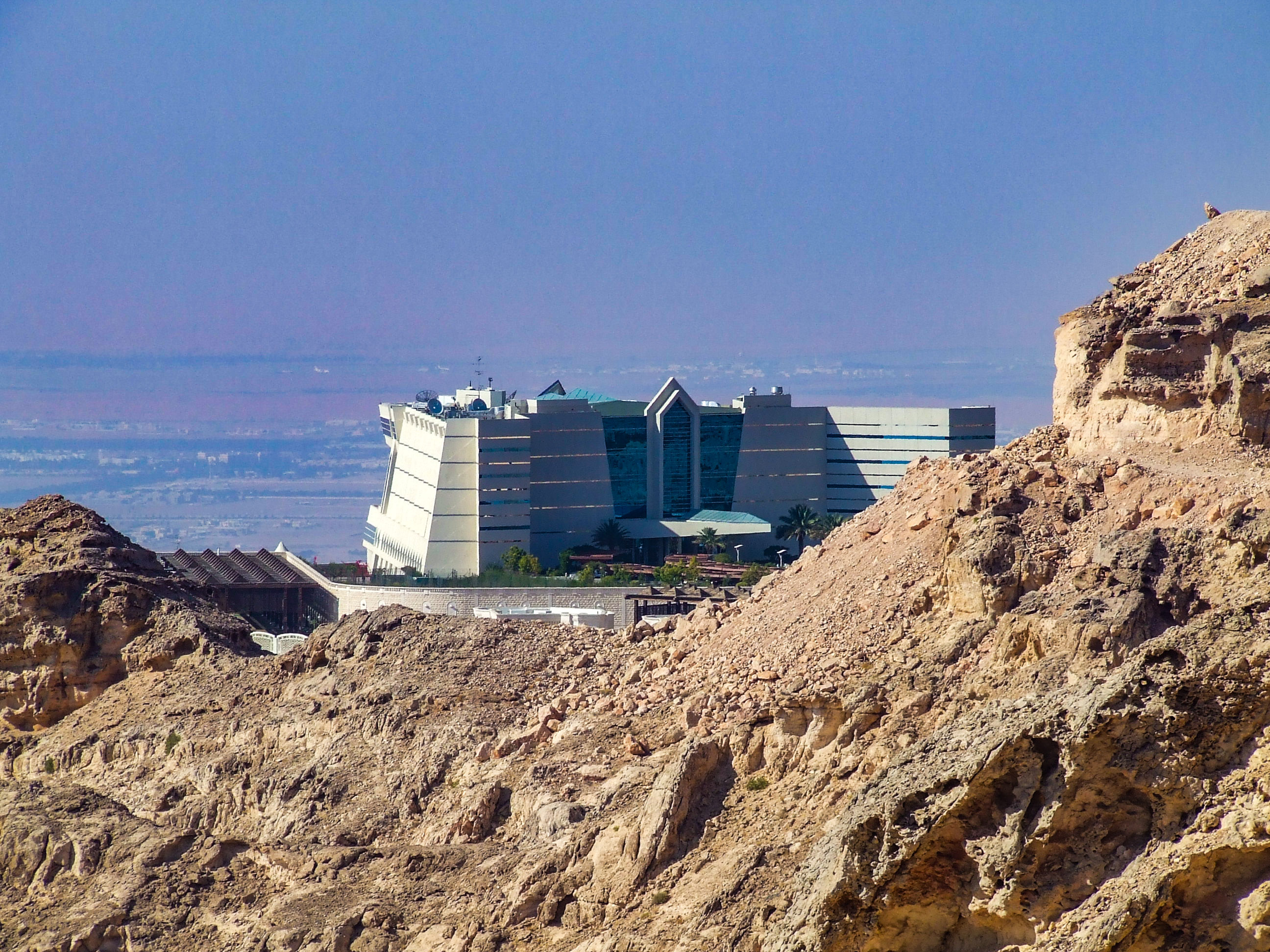 The image depicts a modern building situated on the edge of a rocky cliff, with a vast desert landscape in the background. The building has a unique architectural design with multiple sections and large glass windows. The rocky foreground contrasts sharply with the sleek, contemporary structure. The sky is clear, and the overall scene suggests a location that could be in a desert or arid region.