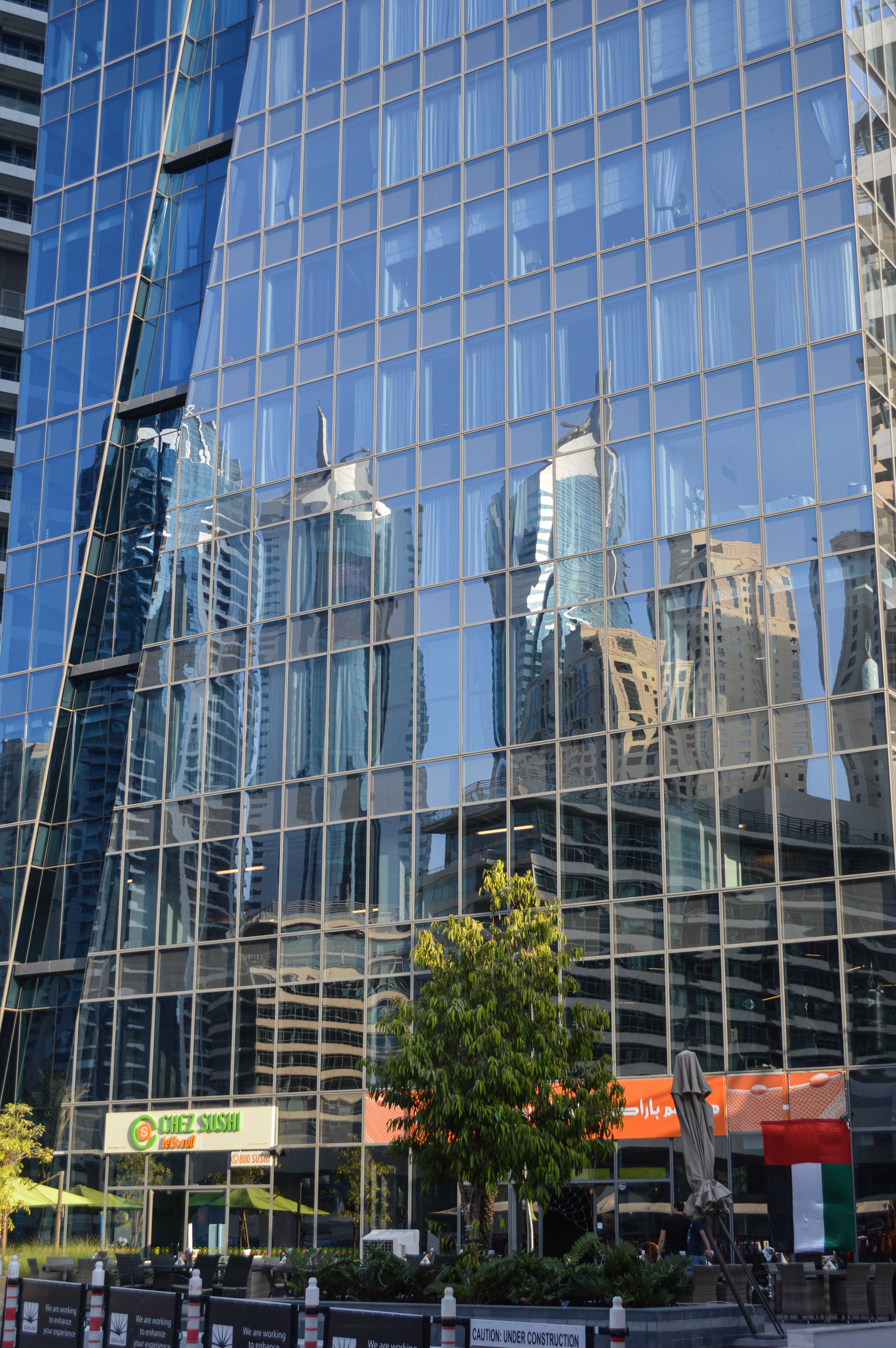 The image depicts a modern urban scene with a large glass building reflecting surrounding skyscrapers and cityscape. The glass facade creates a mirror-like effect, showcasing the architectural diversity of the nearby structures. In front of the building, there is a small outdoor area with trees, plants, and a few signs indicating ongoing construction work. A sign for a restaurant named 'Chel Sushi' is visible at the base of the building, suggesting the presence of commercial establishments within or near the glass building.