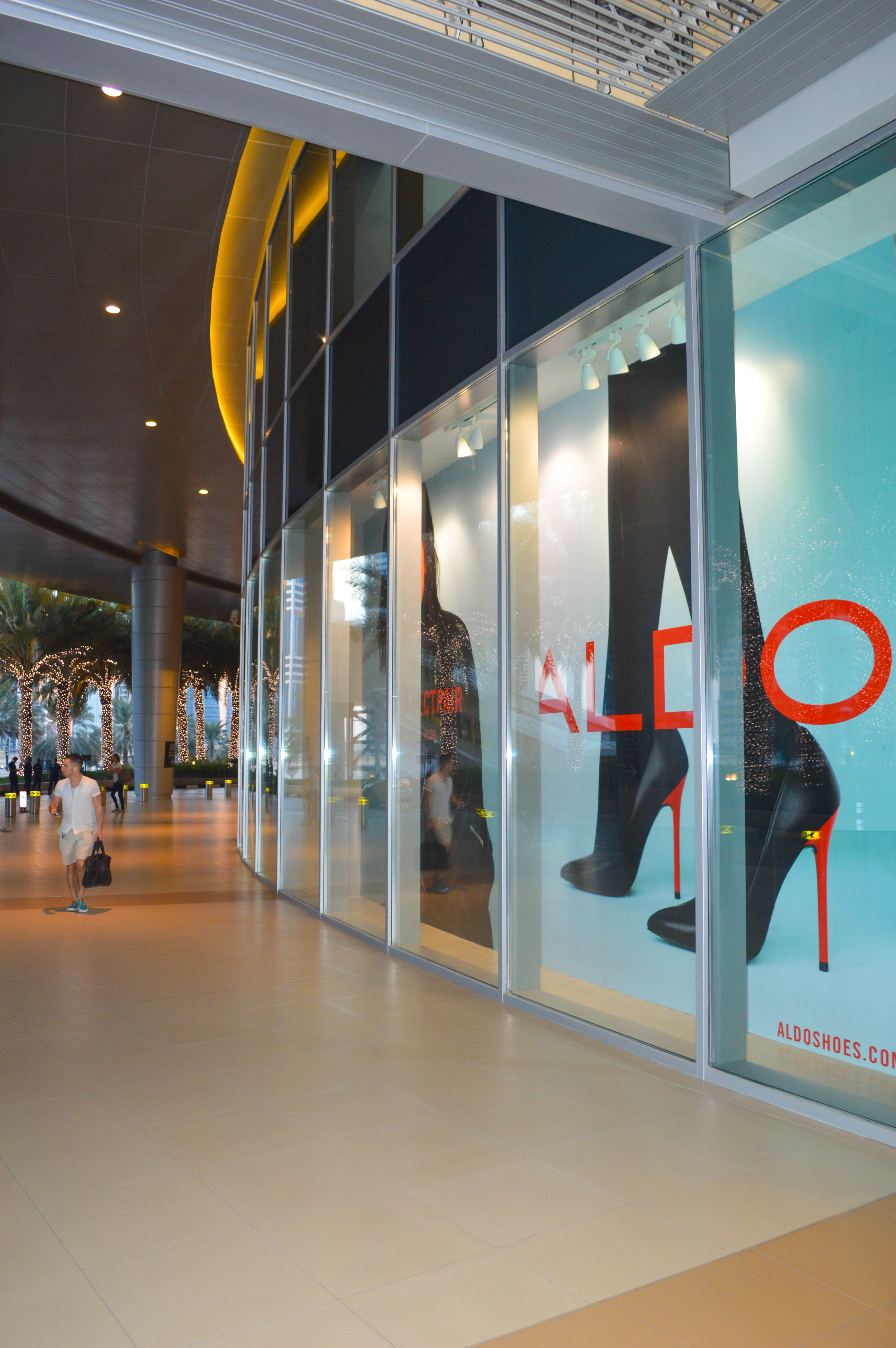 The image depicts a storefront of an Aldo shoe store in a shopping mall. The store features a large glass window display showcasing various styles of women's shoes, including high heels and boots. The interior of the store is well-lit, and the exterior corridor is decorated with festive lights. A man carrying a shopping bag is walking past the store.