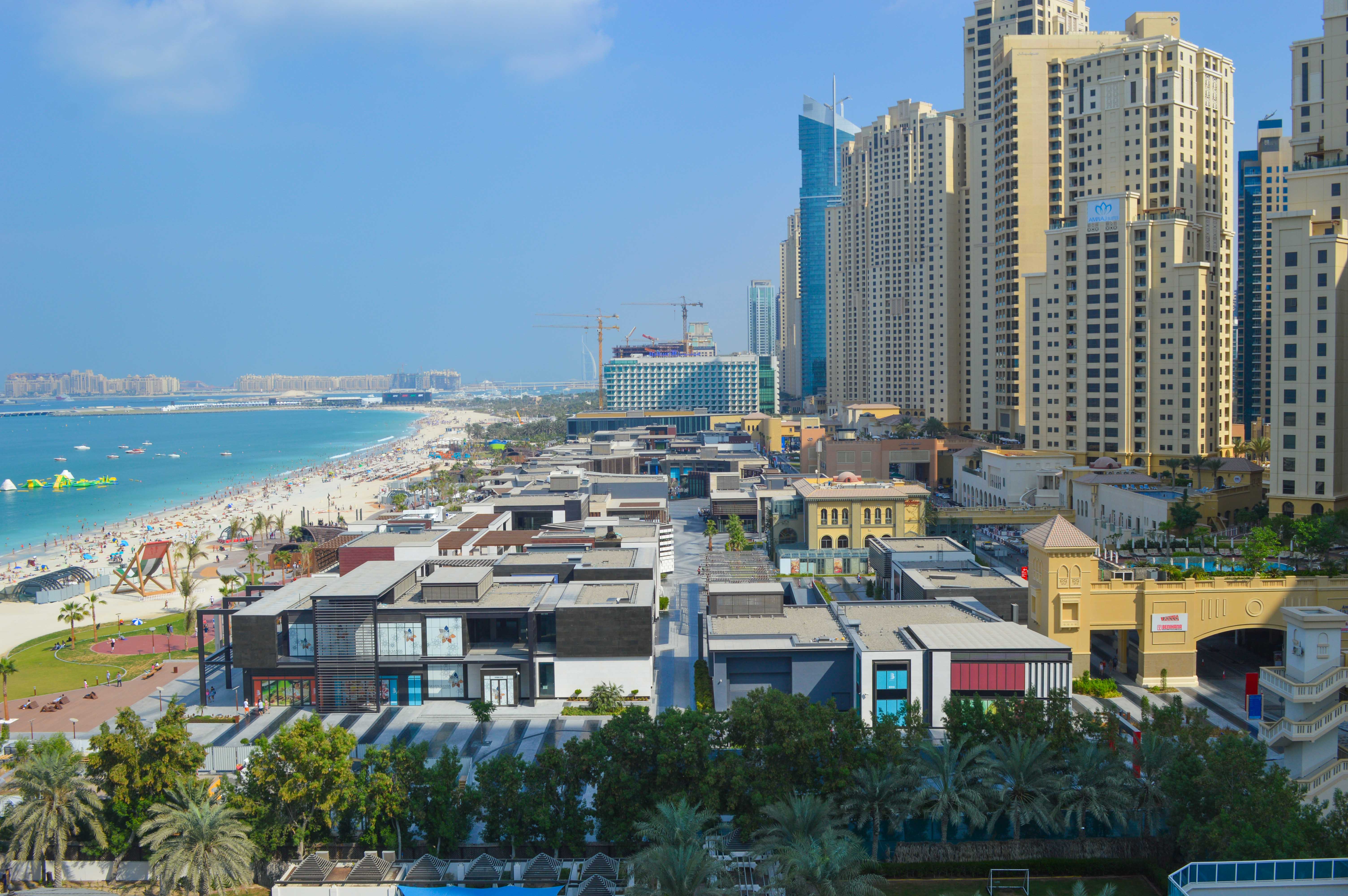 The image depicts a coastal cityscape with tall skyscrapers and modern buildings lining the shore. The beach is crowded with people enjoying the sun and sea. In the foreground, there are various low-rise structures and palm trees, indicating a vibrant urban area with both residential and commercial zones. The overall scene suggests a bustling, developed metropolitan area with a focus on coastal living and tourism.