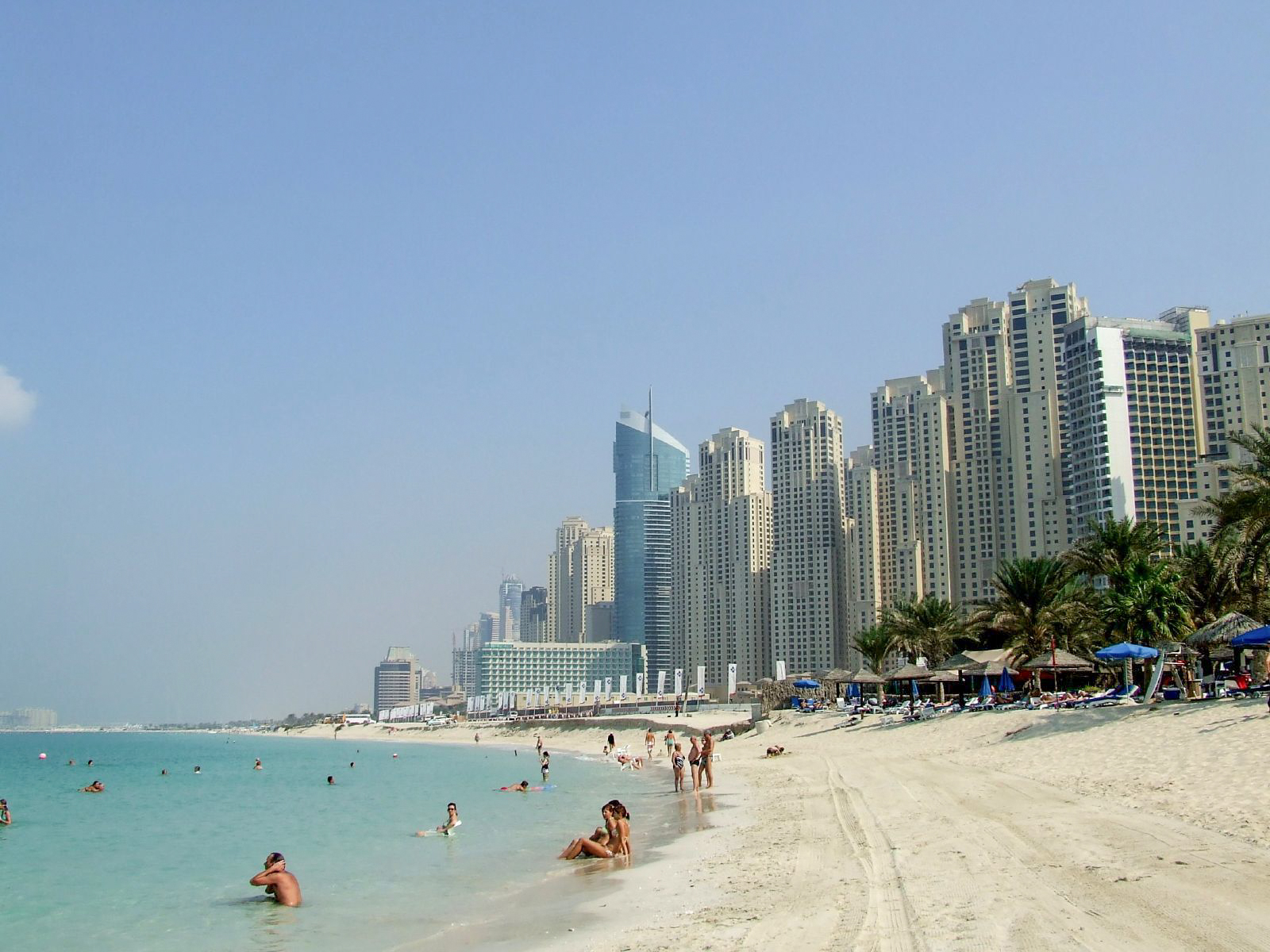 The image depicts a beach scene with people enjoying the water and the sand. In the background, there are numerous tall, modern buildings, indicating an urban coastal area. The sky is clear, and the overall atmosphere appears to be relaxed and sunny.