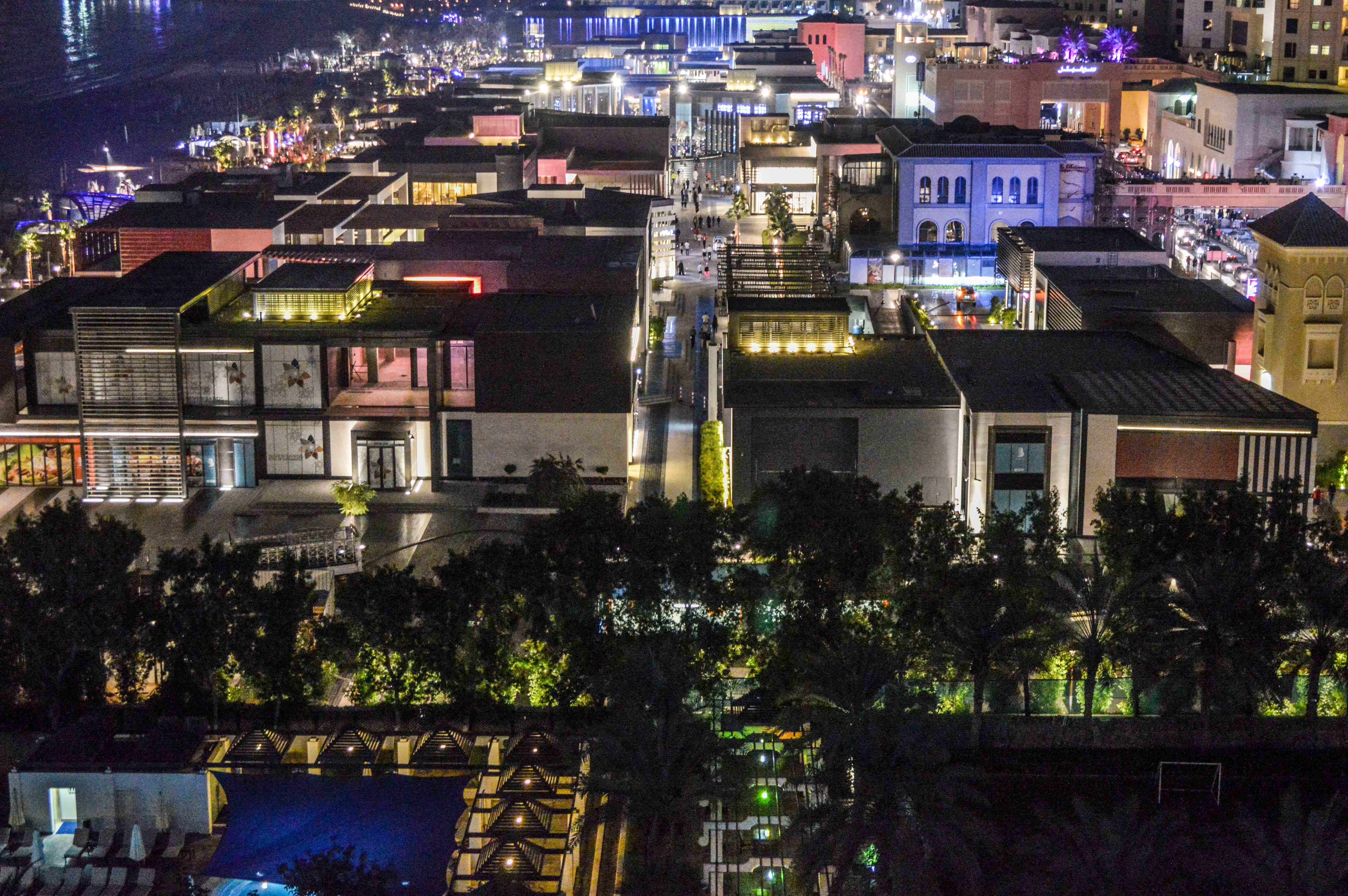 The image depicts a nighttime aerial view of a densely built urban area with well-lit buildings and streets. The architecture varies, with some modern structures and others featuring more traditional designs. The area is vibrant, with numerous lights illuminating the buildings and streets, creating a lively atmosphere. Trees and greenery are visible in the lower part of the image, adding a touch of nature to the urban landscape. The overall scene suggests a bustling, active city environment.