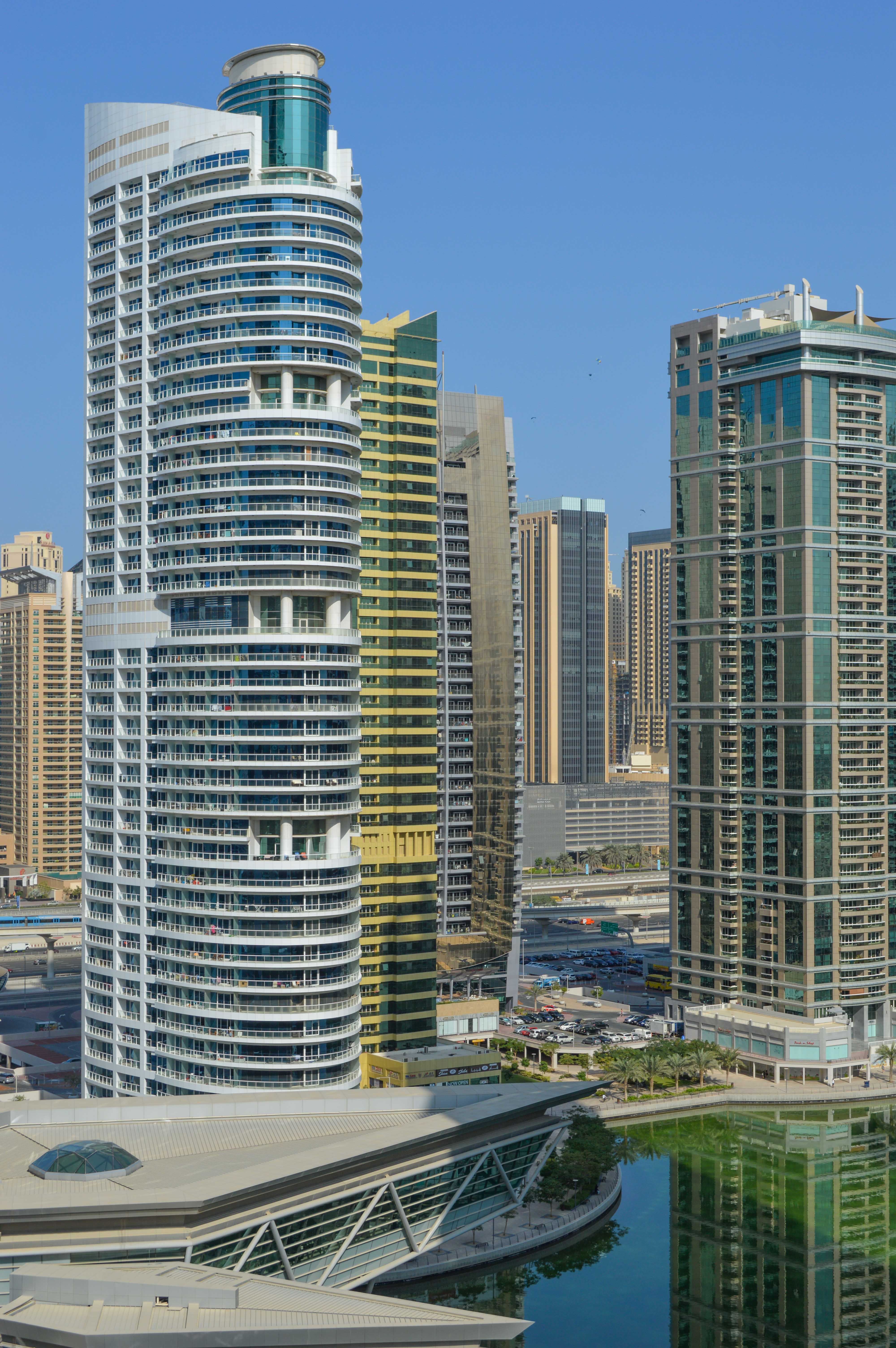 The image depicts a modern cityscape with several high-rise buildings. The architecture is contemporary, featuring a mix of glass and steel structures. The tallest building in the center has a distinctive curved design with multiple balconies. In the foreground, there is a body of water reflecting the buildings, and a unique, angular building with a sloped roof and large glass windows. The sky is clear, indicating a sunny day.