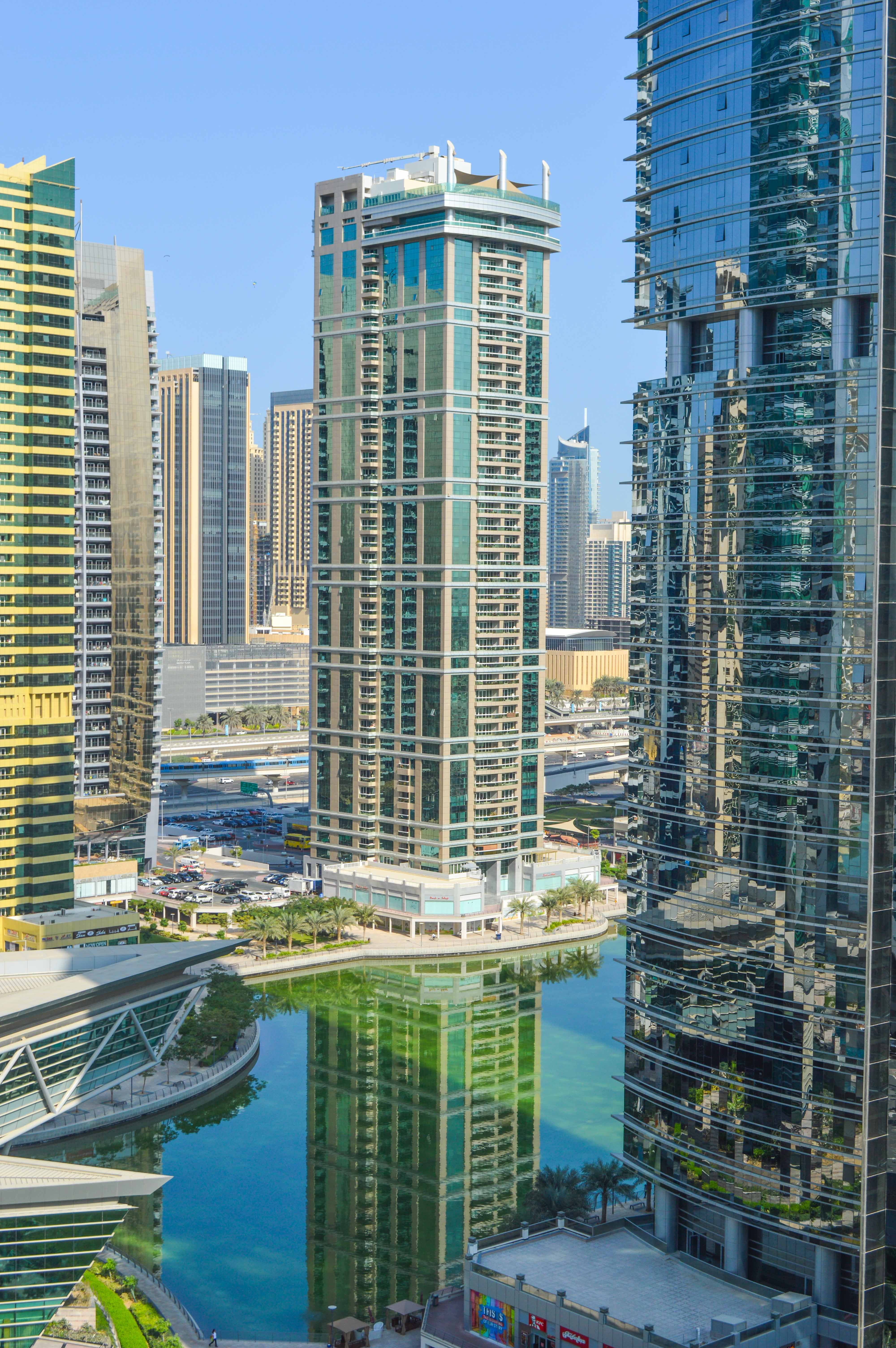 The image depicts a modern urban landscape with tall skyscrapers, reflecting the architectural style of a bustling city. The buildings are predominantly made of glass and steel, showcasing a blend of contemporary design elements. There is a canal or water body running through the center of the image, adding a scenic touch to the urban environment. The area appears to be well-planned with roads, greenery, and pedestrian pathways. The overall scene suggests a vibrant and dynamic cityscape, likely in a major metropolitan area.