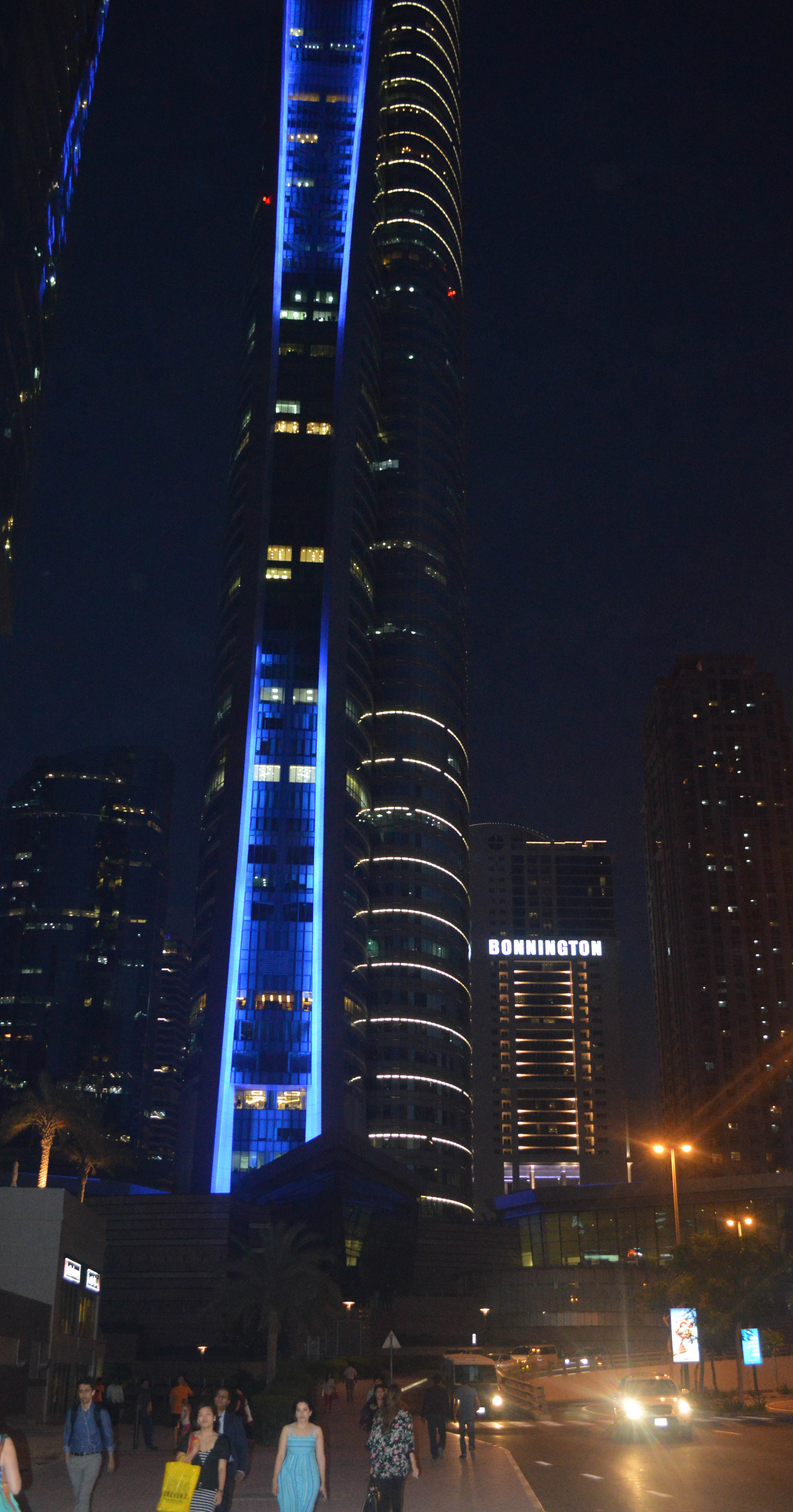 The image depicts a nighttime cityscape featuring tall, illuminated skyscrapers. The central building is prominently lit with blue lights, creating a striking visual effect. The street below is bustling with pedestrians and vehicles, adding to the lively urban atmosphere. The building on the right has the name 'BONNINGTON' illuminated on its facade. The scene captures the essence of a vibrant, modern city at night.