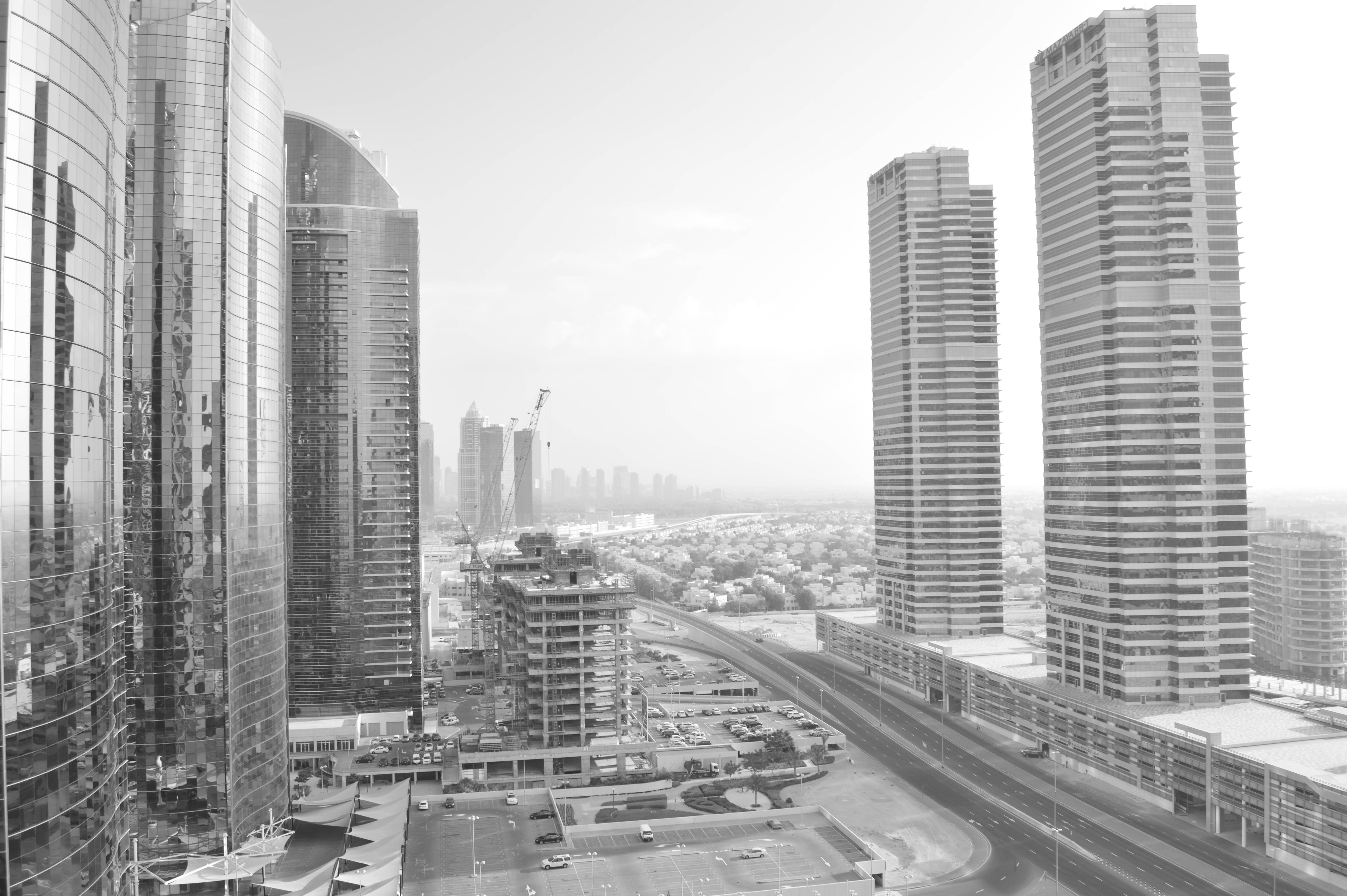The image depicts a black and white photograph of a modern urban landscape featuring several high-rise buildings. The buildings are primarily made of glass and steel, reflecting a contemporary architectural style. The scene includes a mix of completed and under-construction structures, with cranes visible in the background indicating ongoing development. The streets below are relatively empty, suggesting a quiet moment in the city. The overall atmosphere is one of a bustling, developing metropolis with a focus on modern design and infrastructure