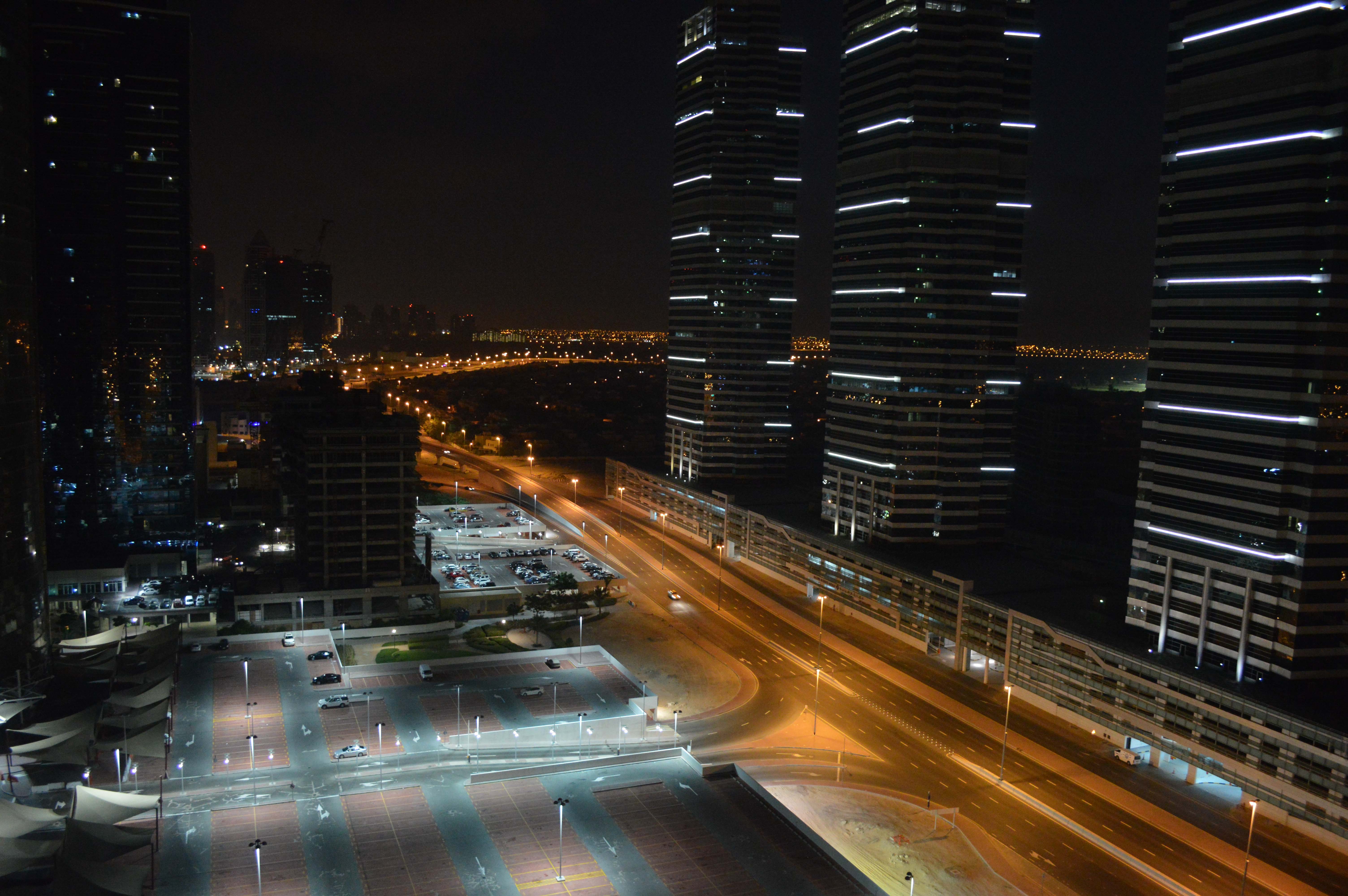 The image depicts a nighttime cityscape featuring tall, illuminated skyscrapers and well-lit roads with minimal traffic. The scene includes a large parking area and a bridge in the distance, with a mix of modern buildings and some construction activity visible in the background.
