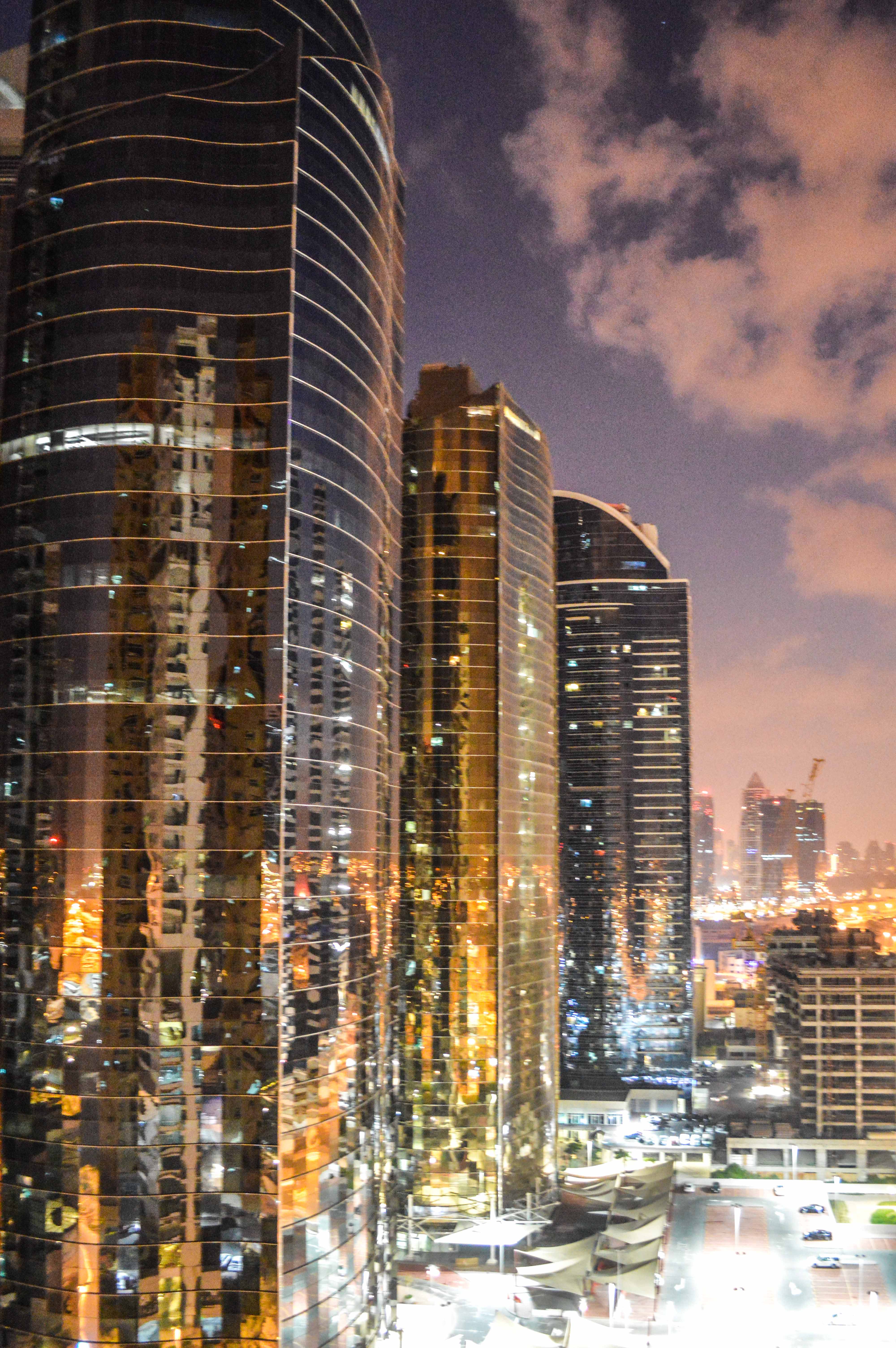 The image depicts a nighttime cityscape featuring several tall, modern skyscrapers with reflective glass facades. The buildings are illuminated by city lights, creating a vibrant and dynamic scene. The sky is partly cloudy, adding depth to the image. The city appears bustling and lively, with a mix of architectural styles and well-lit streets below.