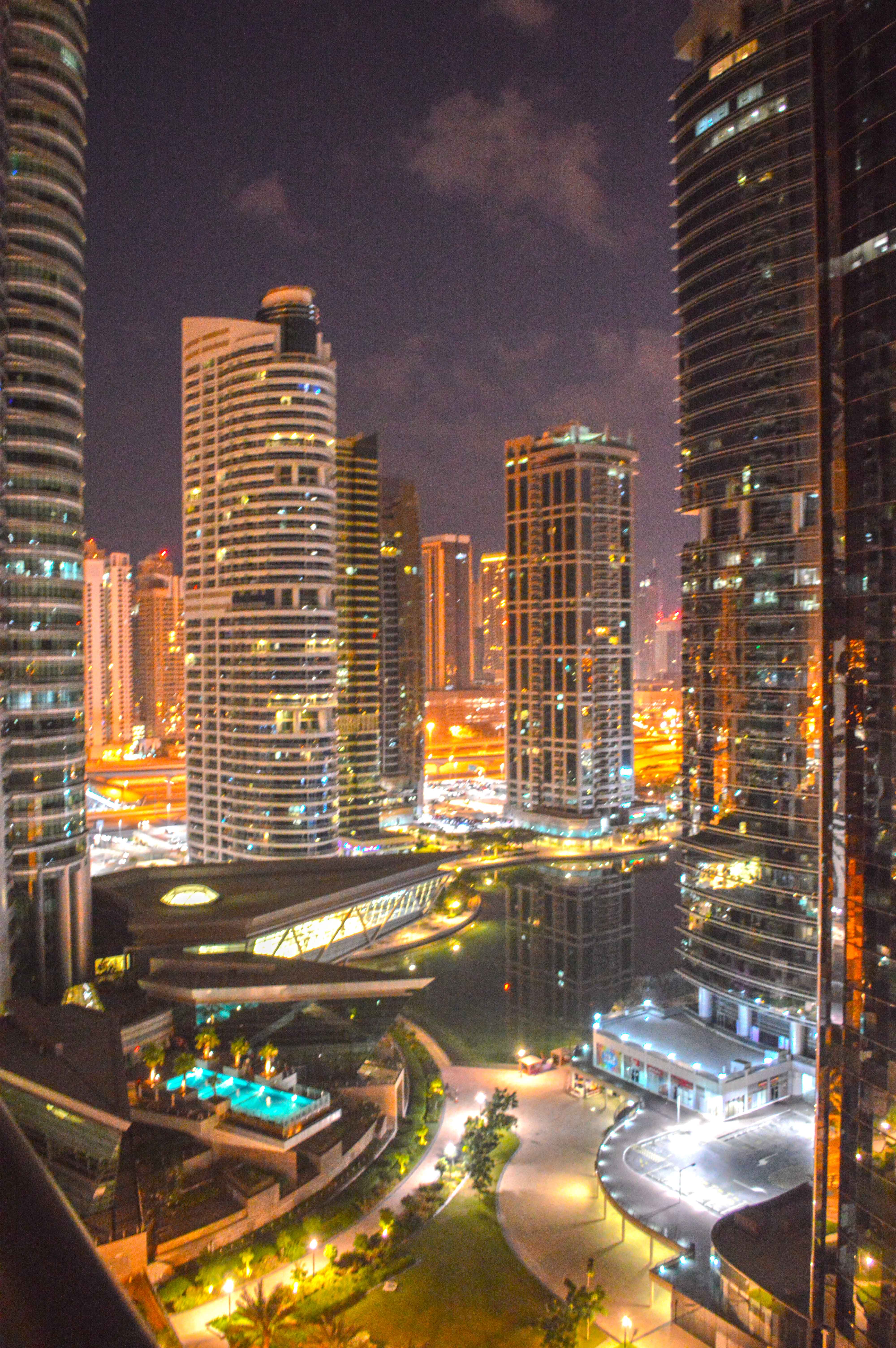 he image depicts a vibrant nighttime cityscape featuring tall, illuminated skyscrapers with a variety of architectural designs. The scene includes well-lit streets with moving vehicles and a modern pedestrian bridge connecting two buildings. In the foreground, there is a landscaped area with pathways, streetlights, and a pool, adding a touch of greenery and recreational space to the urban environment.