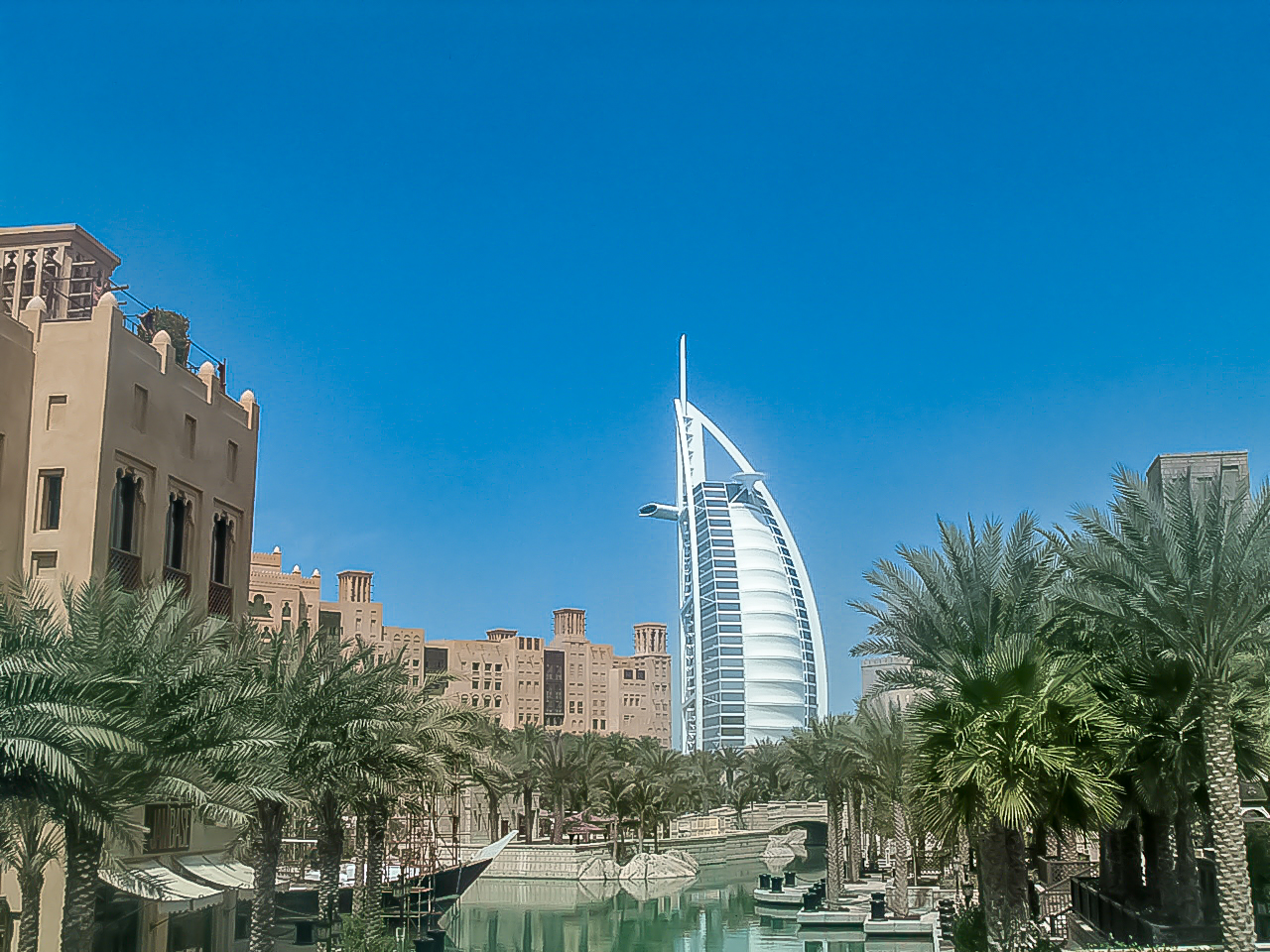 The image showcases a scenic view of the Burj Al Arab, a prominent luxury hotel in Dubai, United Arab Emirates. The hotel's distinctive sail-like shape stands out against the clear blue sky. Surrounding the hotel are lush palm trees and a serene waterway, adding to the picturesque setting. The architecture in the foreground features traditional Middle Eastern design elements, creating a blend of modern and traditional aesthetics. The overall scene highlights the unique and luxurious atmosphere of Dubai.