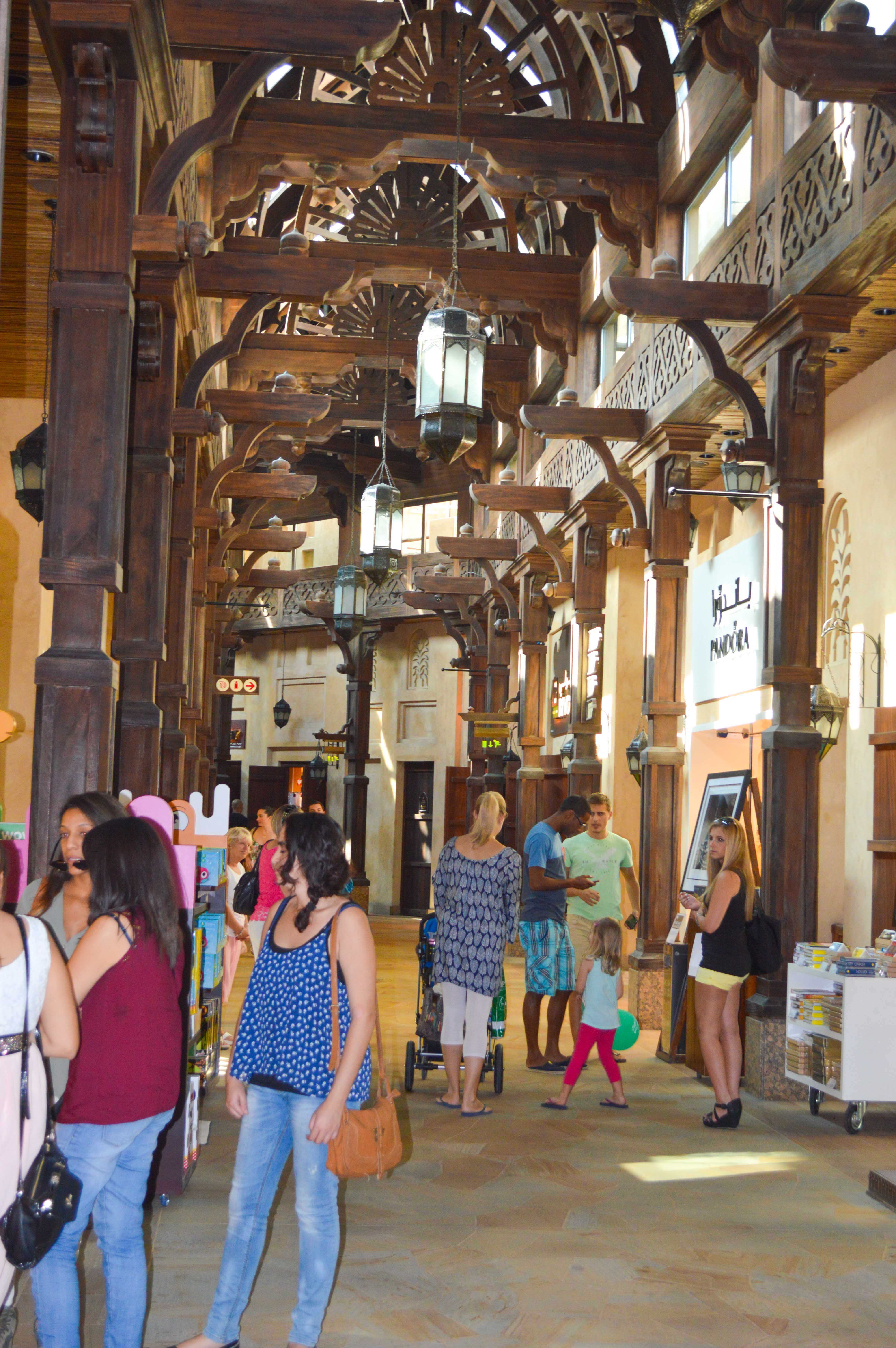 The image depicts a bustling indoor market or shopping area with a distinctive architectural style featuring wooden beams and lanterns. Several people are seen walking, shopping, and interacting with each other. The area appears to be well-lit with natural light streaming in from the sides. Various stalls or shops are visible, selling different items. The overall atmosphere seems lively and vibrant.