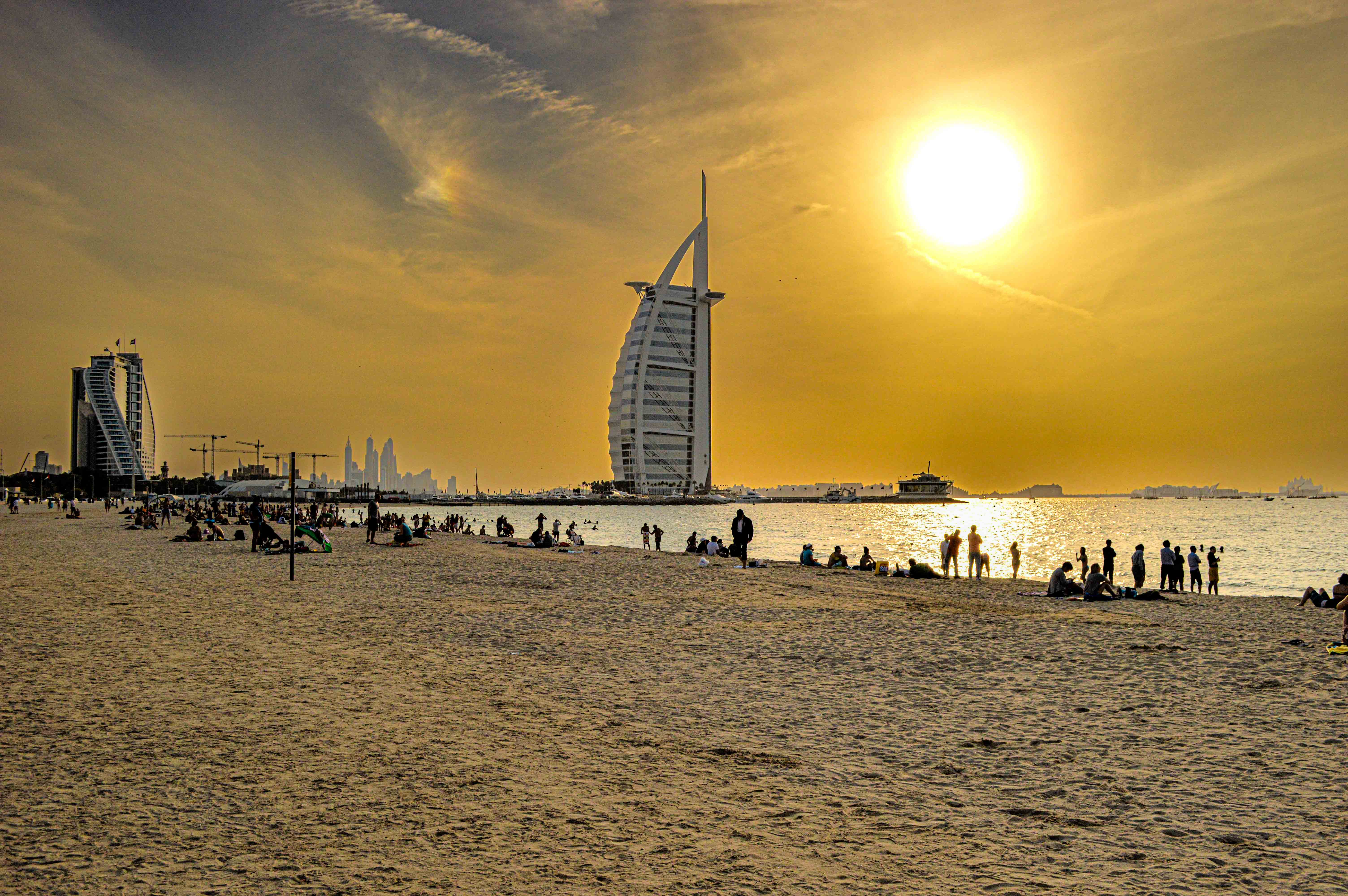 The image depicts a beach scene at sunset with numerous people enjoying the beach. In the background, there are iconic buildings, including the distinctive Burj Al Arab, a luxury hotel in Dubai. The sky is filled with warm hues of orange and yellow, casting a golden glow over the scene. The beach is bustling with activity, with people walking, sitting, and relaxing on the sand. The overall atmosphere is lively and serene, capturing a typical evening at a popular beach destination.