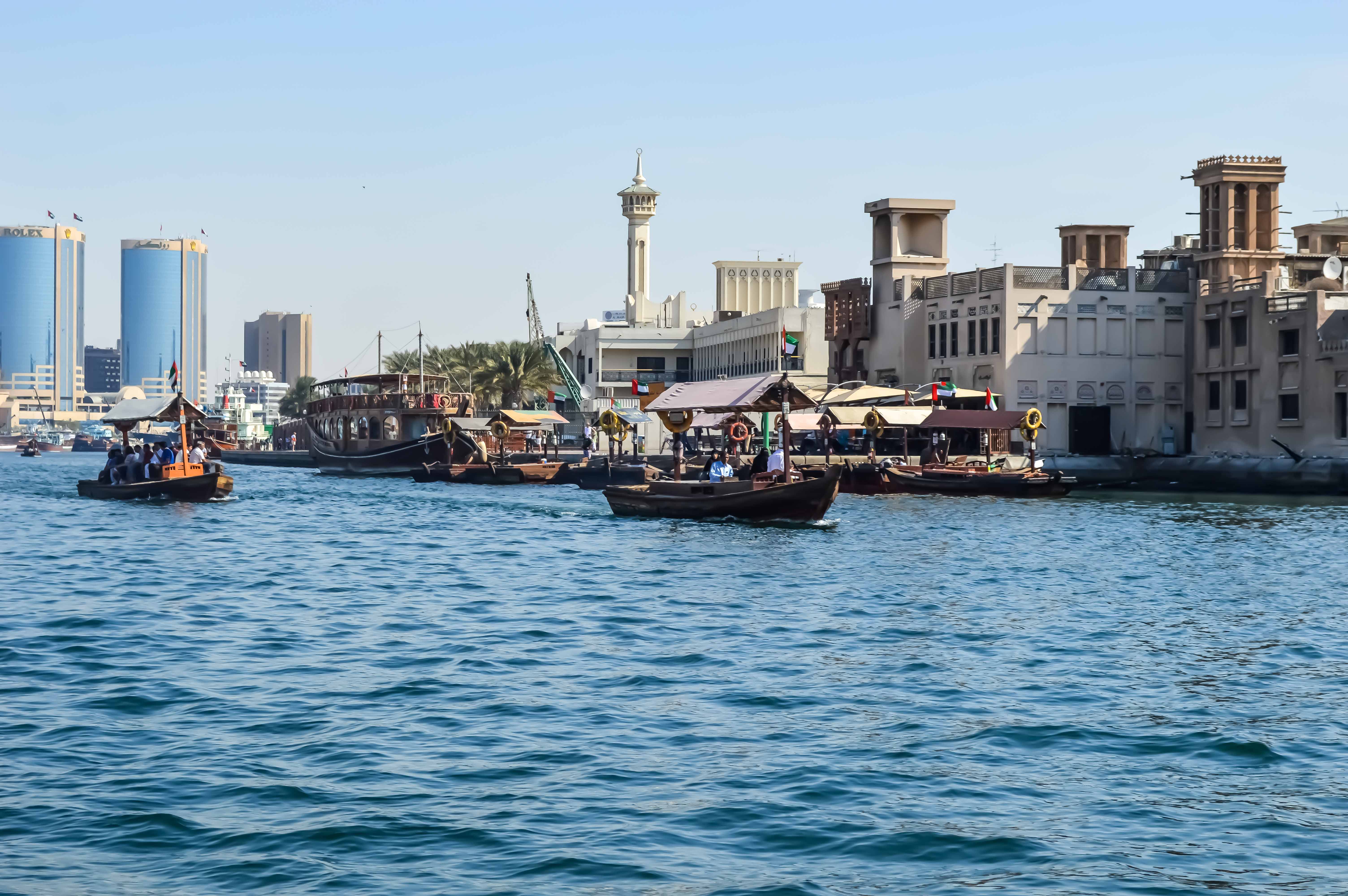 The image depicts a bustling waterfront scene in a modern city with traditional elements. Several small boats are navigating the water, while larger vessels are docked along the shore. The backdrop features a mix of contemporary high-rise buildings and traditional architecture, including a prominent minaret. The scene suggests a blend of cultural heritage and modern development, likely in a Middle Eastern city.