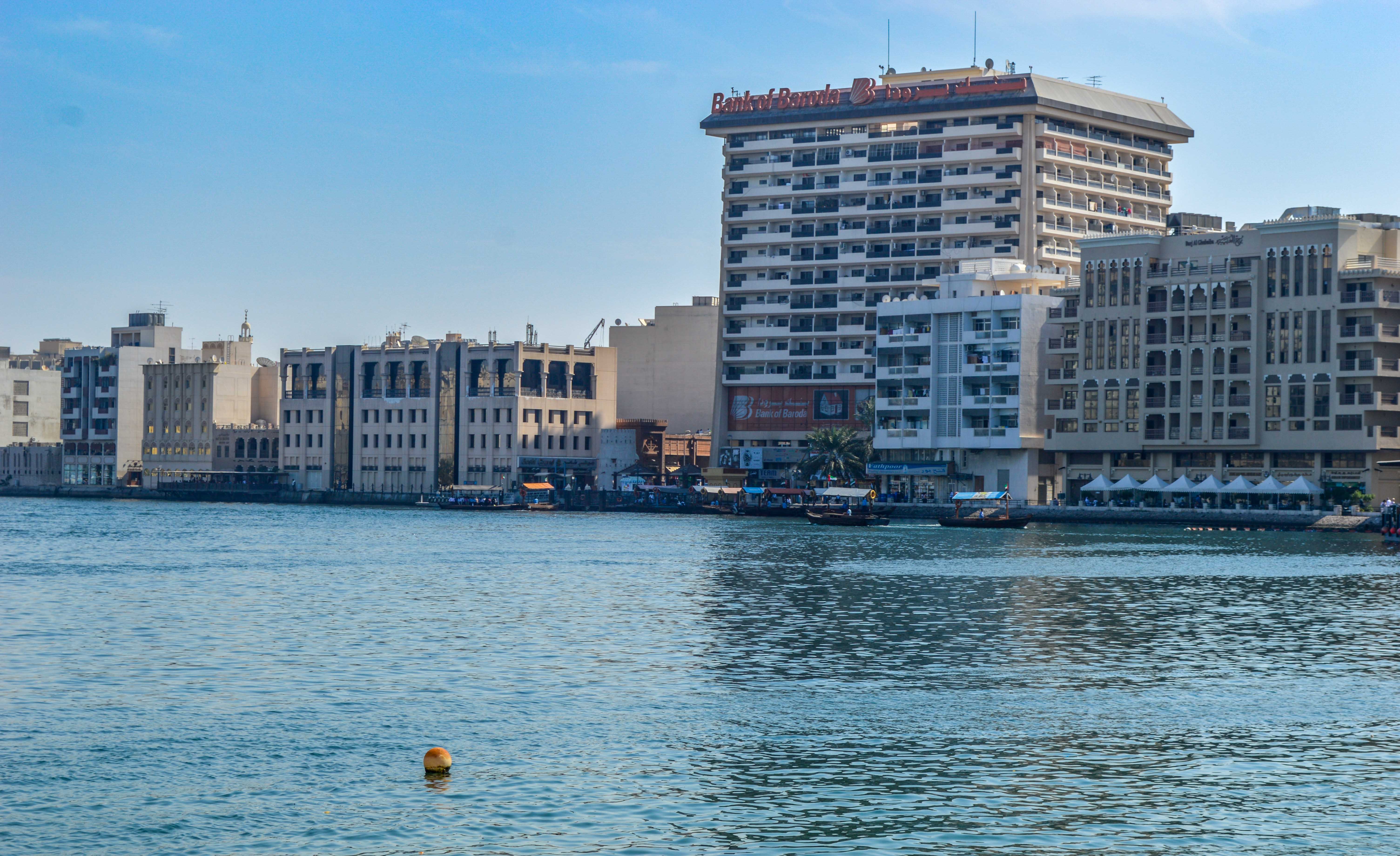 The image depicts a waterfront scene with modern buildings, including a prominent high-rise labeled 'Bank of Baroda.' The buildings are situated along the shore, with several boats and a dock area visible in the foreground. The water is calm, and the sky is clear, suggesting a serene and sunny day