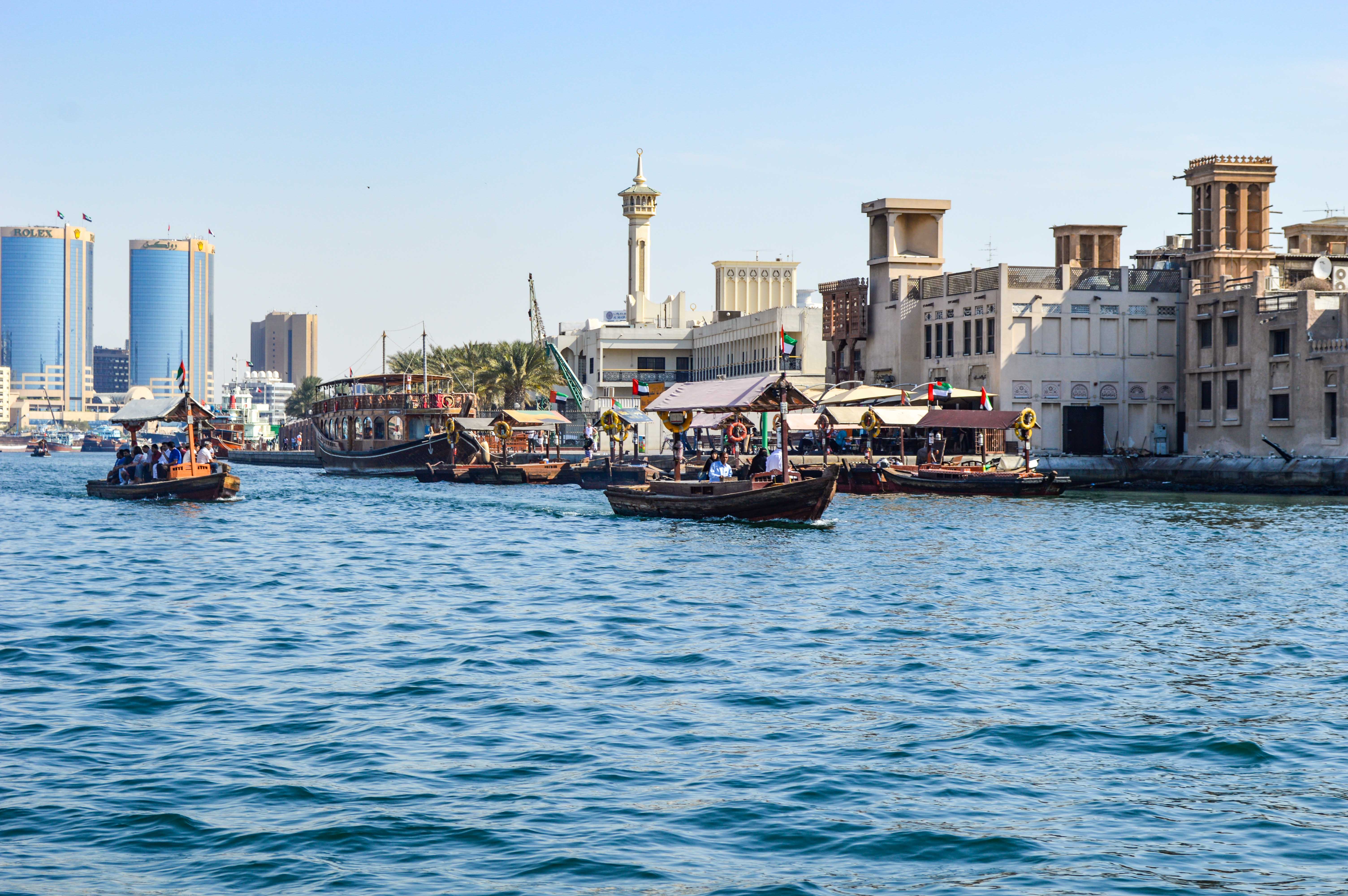 The image depicts a vibrant waterfront scene in a modern city with traditional elements. Several small boats are on the water, with people enjoying a ride. In the background, there is a mix of modern skyscrapers and traditional buildings, showcasing a blend of old and new architecture. The scene is lively, with various activities happening along the waterfront, including what appears to be a docked traditional boat serving as a restaurant or cafe. The overall atmosphere is bustling and dynamic, reflecting a city that embraces both its heritage and contemporary development.