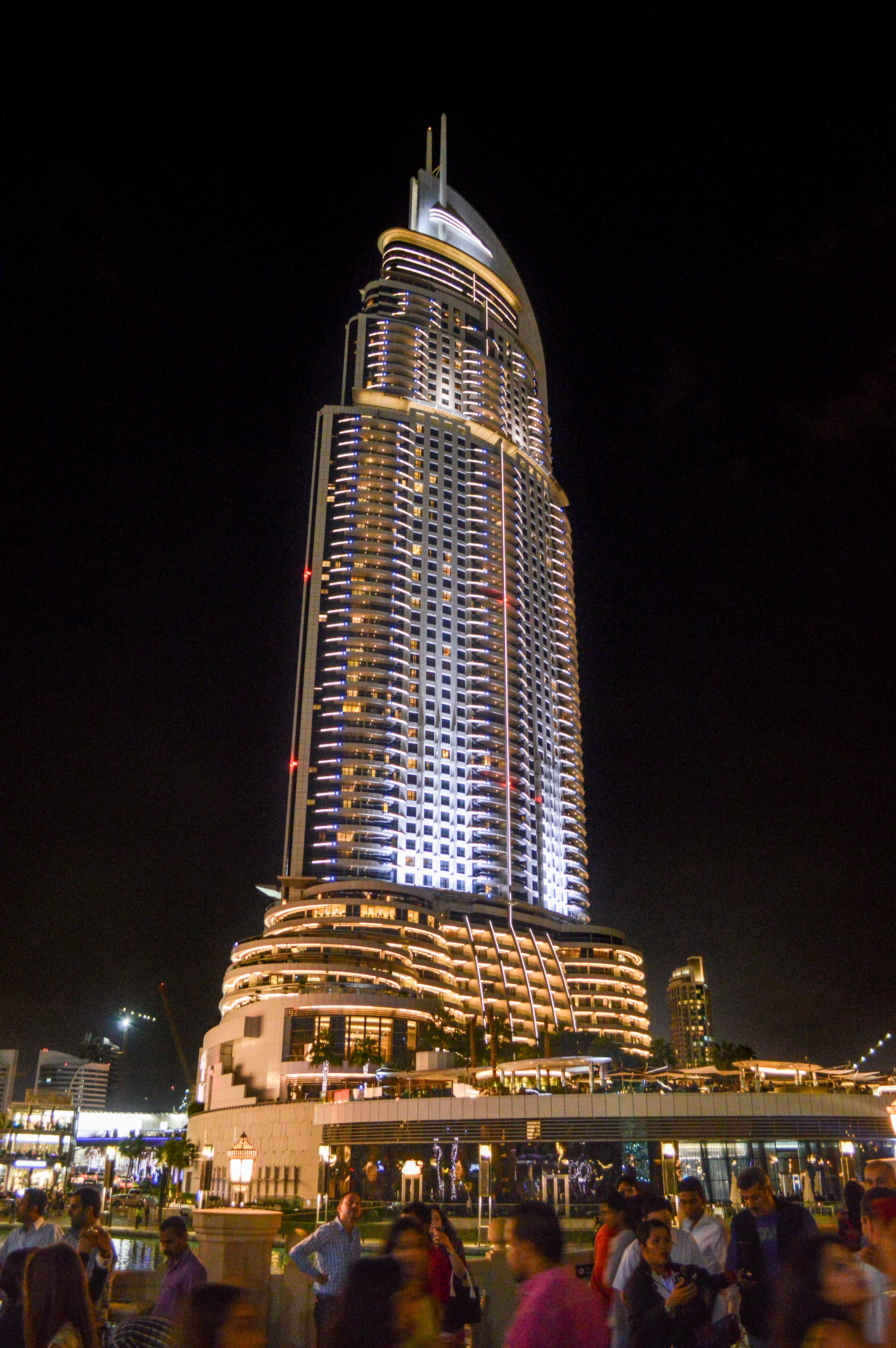 The image depicts a tall, illuminated skyscraper at night with a distinctive curved shape and multiple balconies. The building is surrounded by a bustling crowd of people at its base, indicating a lively urban environment. The structure is well-lit with a combination of white and blue lights, creating a striking visual contrast against the dark night sky. The lower levels of the building feature extensive outdoor seating and dining areas, suggesting a vibrant social scene. The overall atmosphere is energetic and modern, characteristic of a popular city landmark.