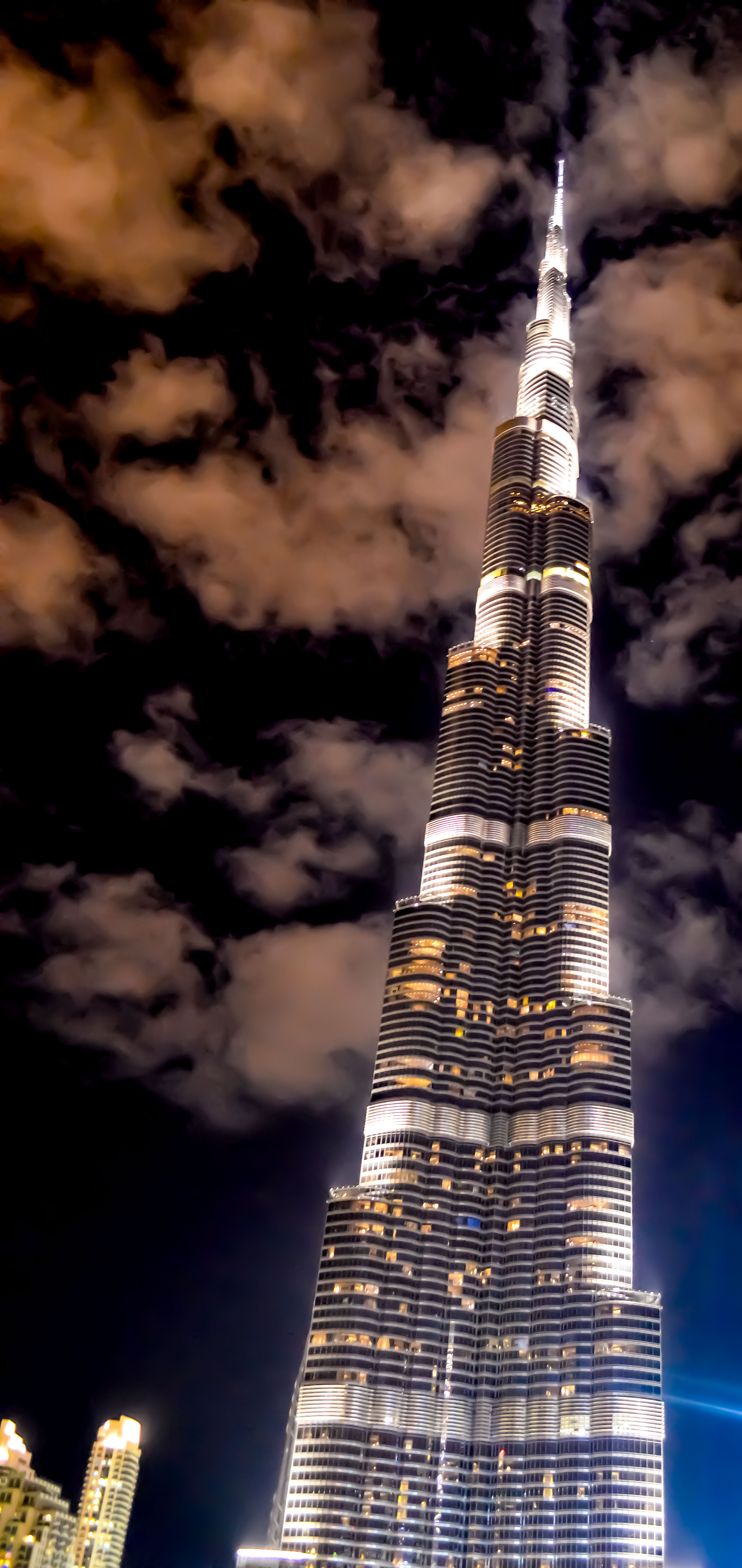 The image depicts the Burj Khalifa, the tallest building in the world, located in Dubai, United Arab Emirates. It is shown at night with its lights illuminated, standing tall against a backdrop of clouds.