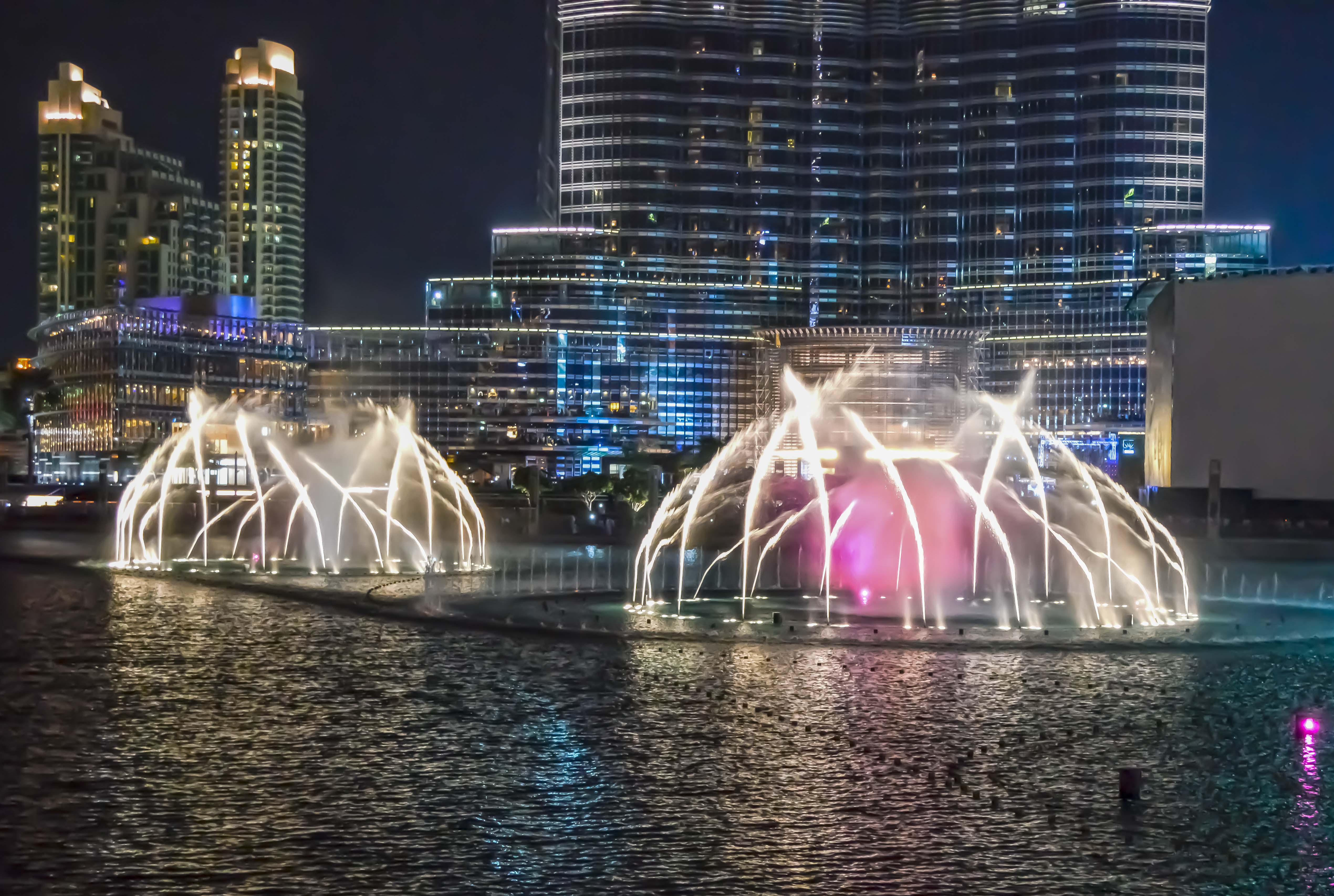 The image depicts a nighttime scene of the Dubai Fountain show in Dubai, UAE. The fountain jets are illuminated with colorful lights, creating a spectacular display against the backdrop of tall, modern buildings. The water movements are synchronized with the lights, producing an enchanting visual effect. The surrounding skyscrapers are brightly lit, adding to the overall vibrancy of the scene. The reflection of the fountains and lights can be seen in the water below, enhancing the visual appeal.