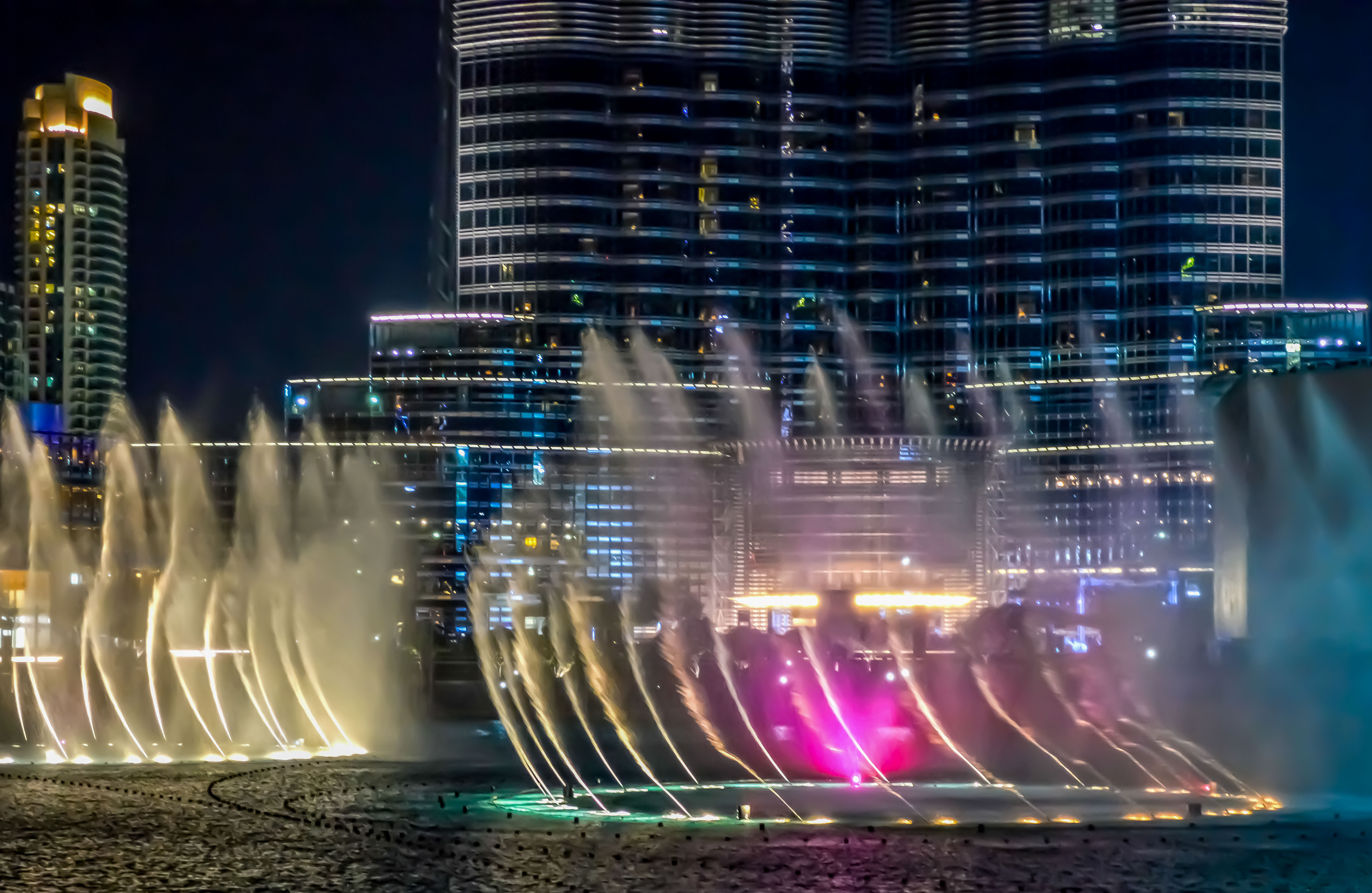 The image depicts a night scene of a modern cityscape with tall, illuminated skyscrapers. In the foreground, there is a dynamic fountain show with water jets creating colorful, arched patterns. The buildings are lit up with various lights, adding to the vibrant atmosphere. The photo captures the essence of urban life and architectural beauty, highlighting the contrast between the static structures and the moving water fountains.
