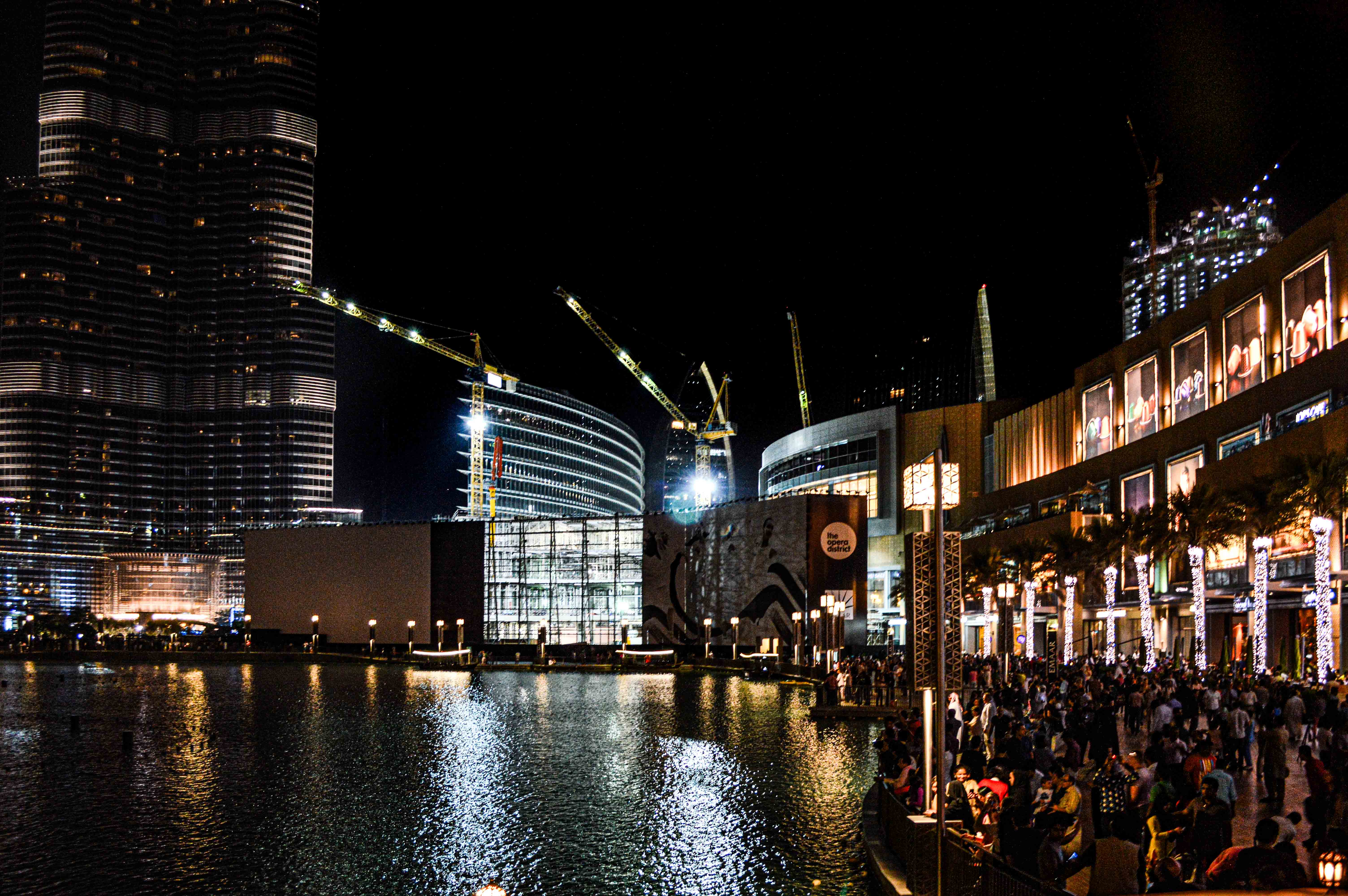 The image depicts a vibrant night scene in a modern urban area, likely a shopping district. Tall, illuminated skyscrapers and contemporary buildings surround a large water feature. Construction cranes indicate ongoing development. The area is bustling with people, suggesting it is a popular destination. The overall atmosphere is lively and dynamic, showcasing the blend of commerce, architecture, and social activity.