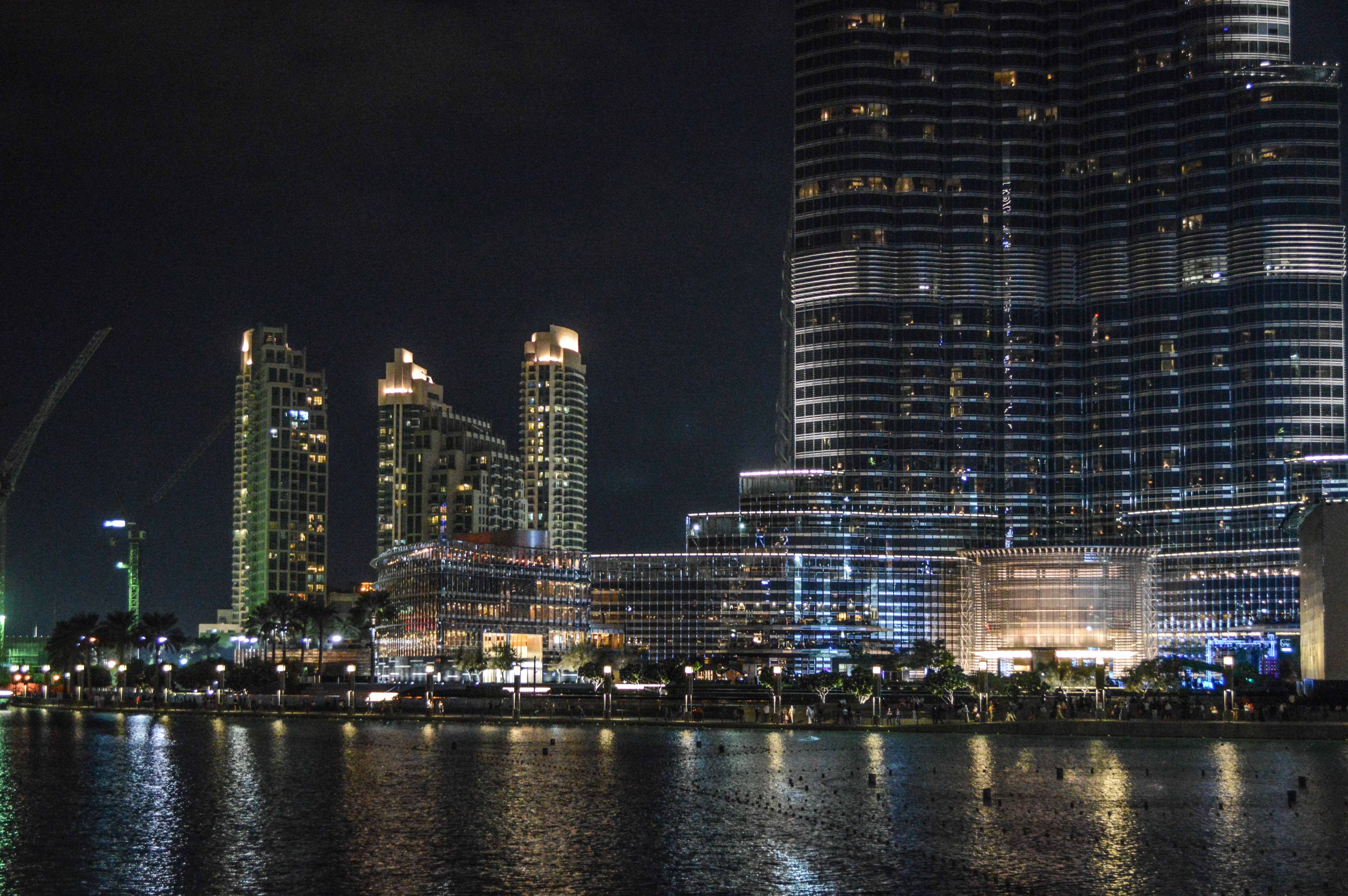 The image depicts a nighttime cityscape featuring modern skyscrapers and high-rise buildings illuminated with various lights. The tallest building on the right is the Burj Khalifa, a prominent landmark in Dubai. The scene includes a body of water in the foreground, reflecting the lights from the buildings. The area appears to be bustling with activity, indicating a lively urban environment.