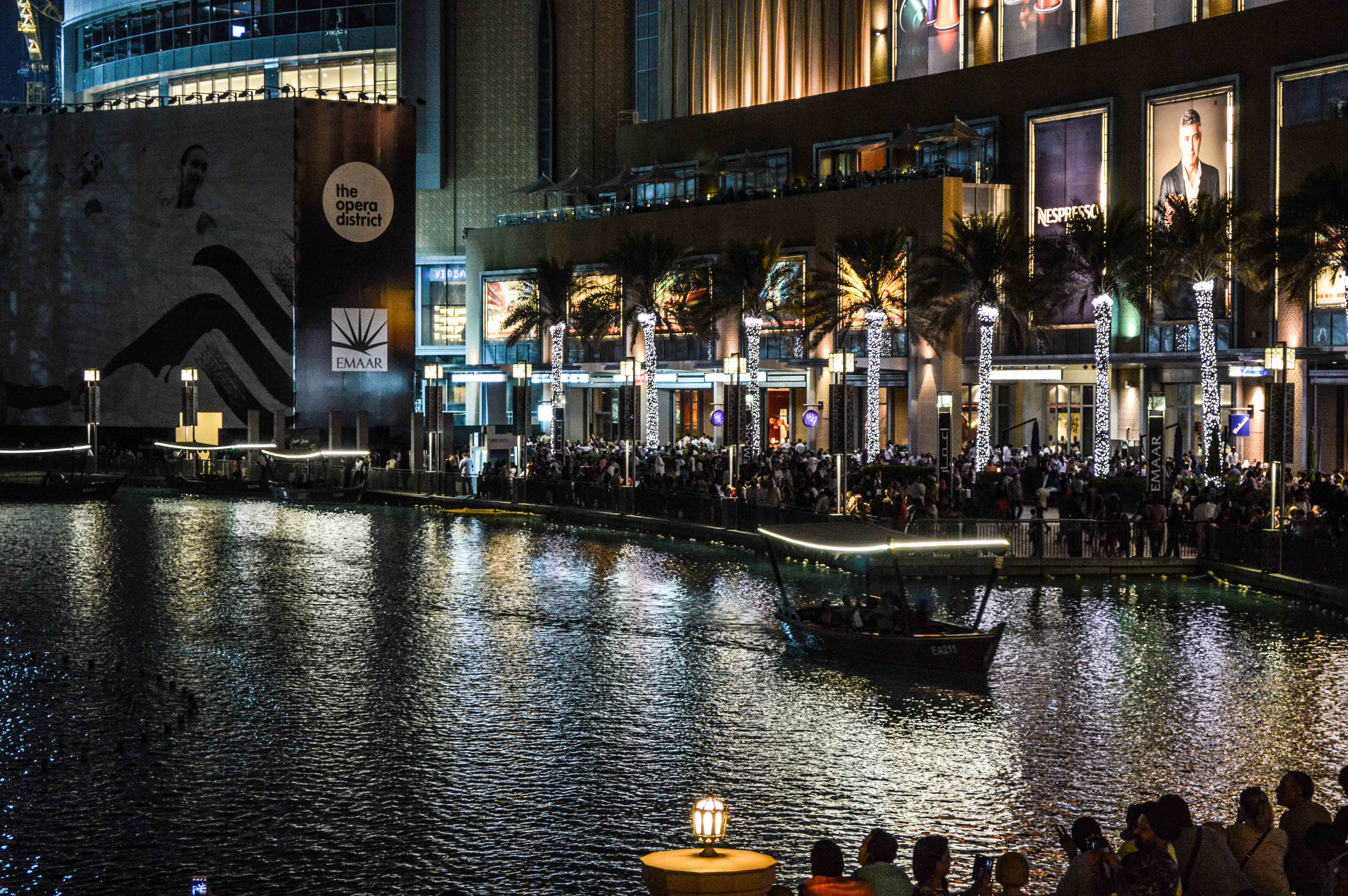 The image depicts a vibrant night scene at The Opera District, a part of EMAAR. The area is bustling with people, and there is a canal with a boat on it. The buildings are illuminated with lights, and there are palm trees and decorative lights along the walkway. The atmosphere appears lively and festive, with numerous individuals gathered along the waterfront.